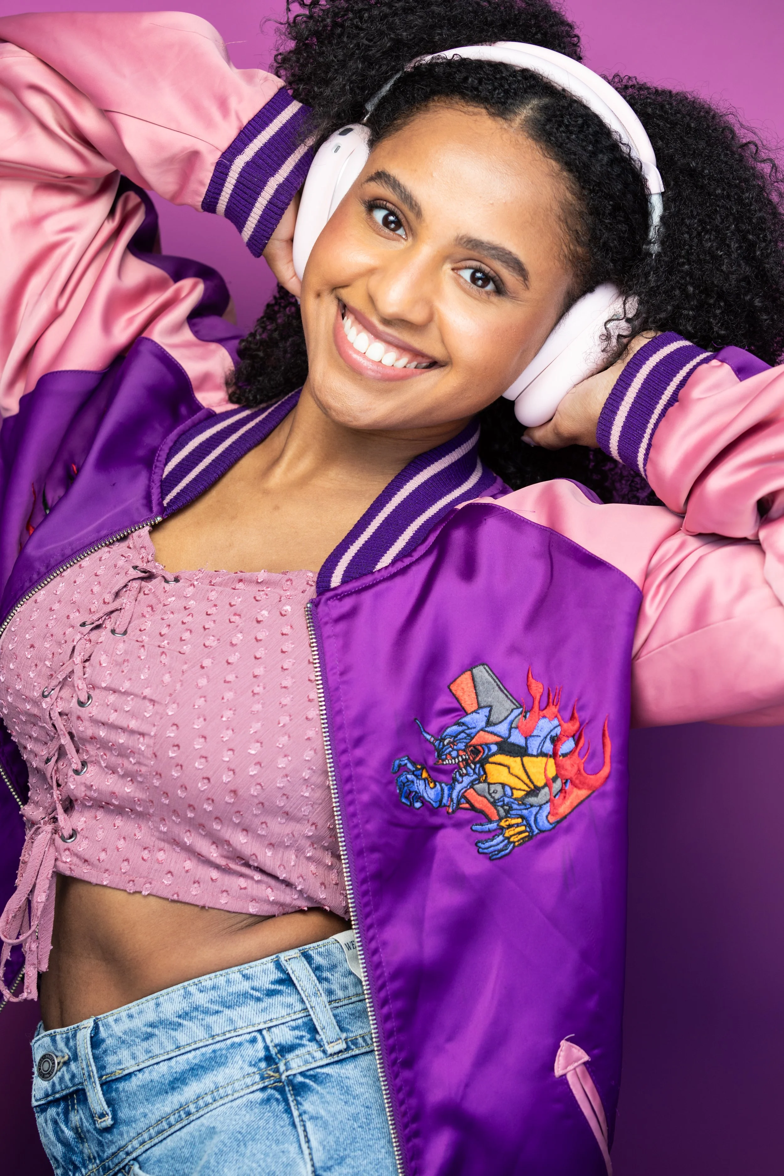 A young woman smiling and wearing white headphones, a purple and pink varsity jacket, a pink textured crop top, and blue jeans, standing in front of a purple background.
