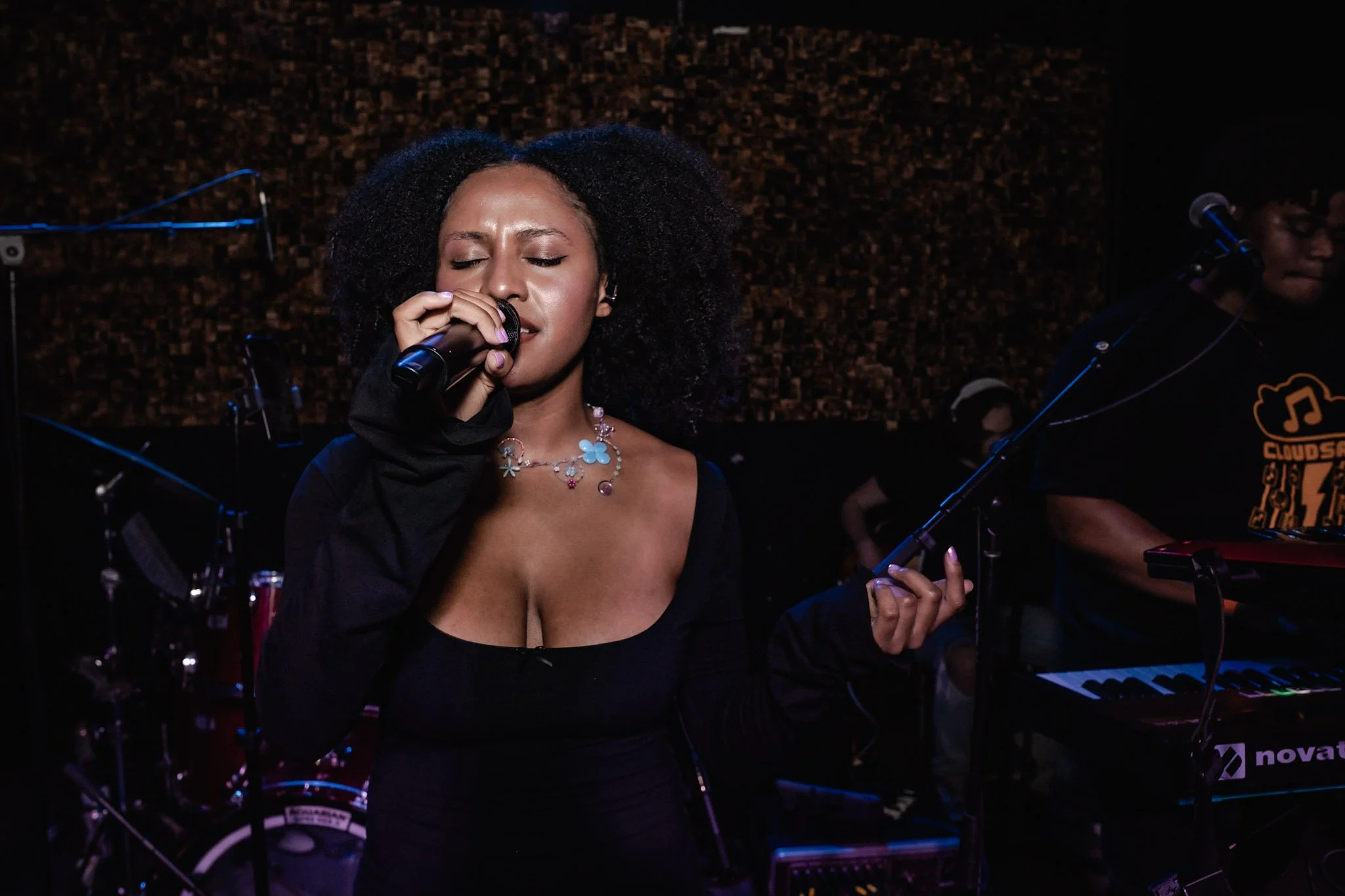 A woman passionately singing into a microphone during a live music performance, with a keyboard player visible in the background.