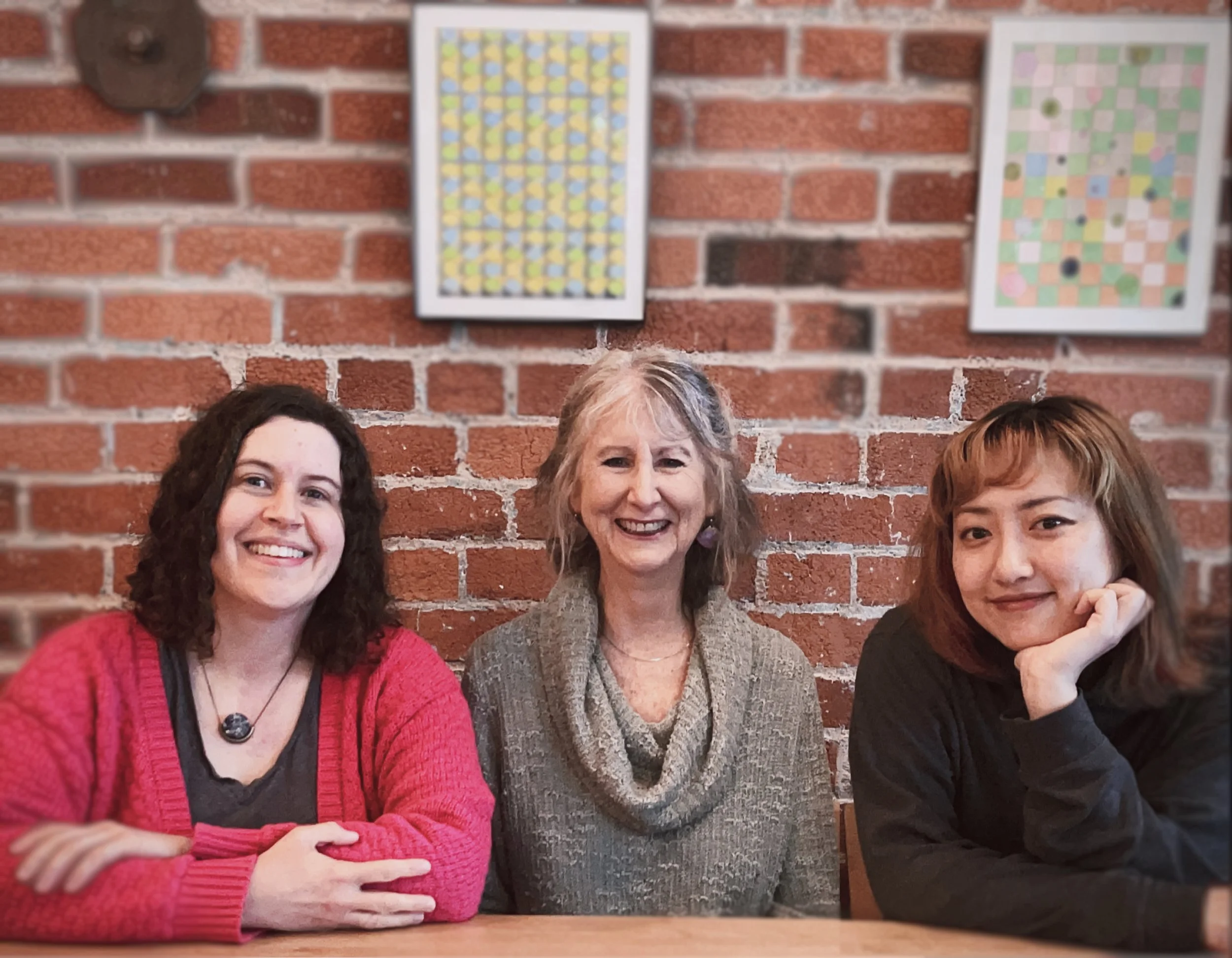 Three smiling women posing together in front of a brick wall with framed artwork.