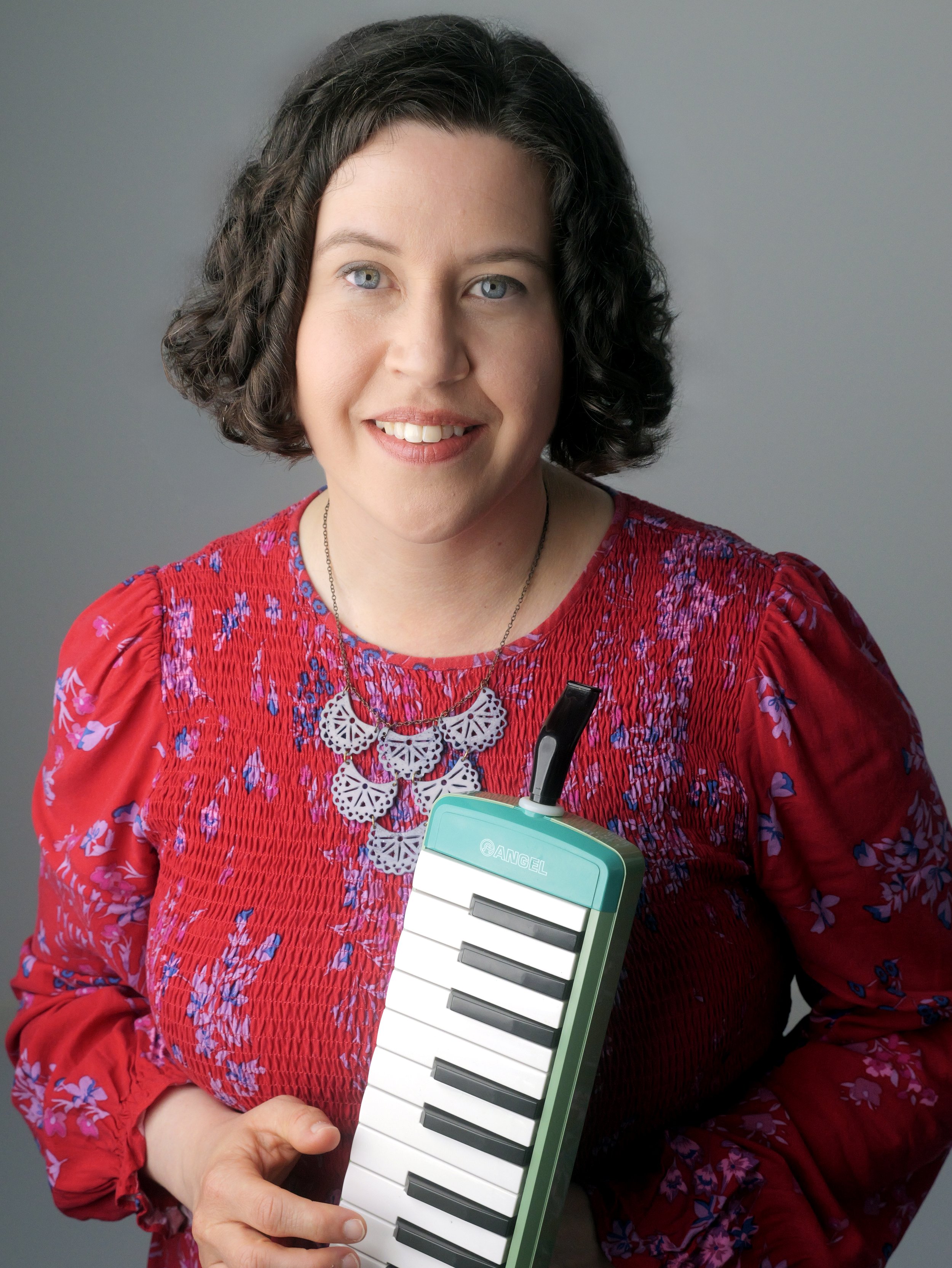 Smiling woman with curly dark hair holding a turquoise-and-white melodica in front of a gray background