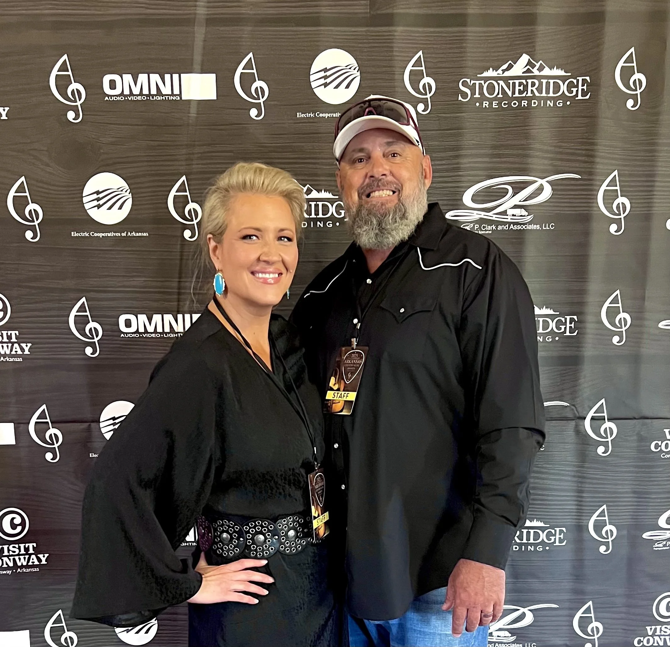 Two people smiling in front of a backdrop with music notes and logos, one wearing a white hat and sunglasses, the other with short blonde hair and turquoise earrings.