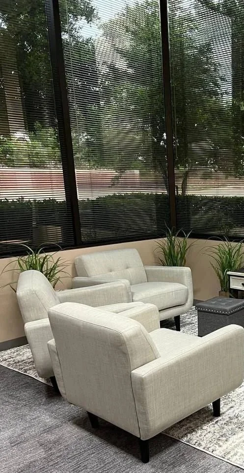 JW Counseling TX therapy room with two beige armchairs and a beige loveseat, a black ottoman, potted plants, and large black-framed windows with blinds showing trees outside.