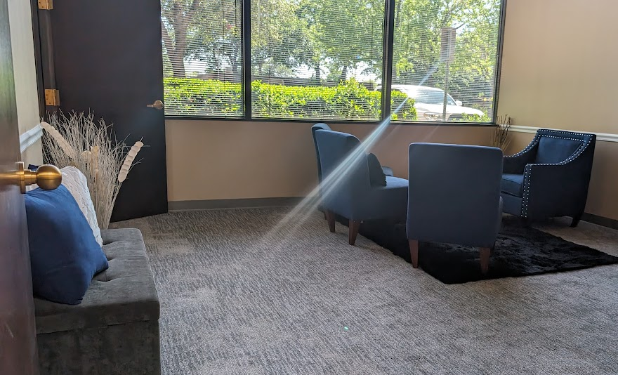 JW Counseling TX therapy room with blue armchairs, a gray sofa with pillows, a black rug, large windows with a view of outdoor greenery, and a door on the left side.