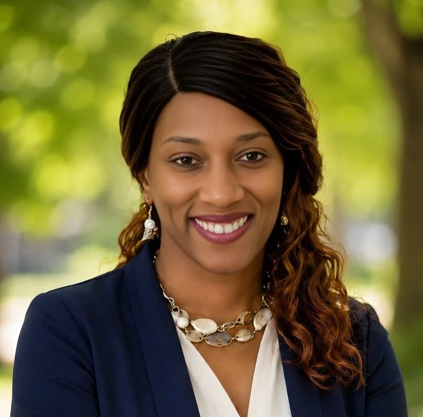 Julie "Jaye" Williams, wearing a navy blazer, white blouse, necklace, and earrings, smiling outdoors with green trees in the background.