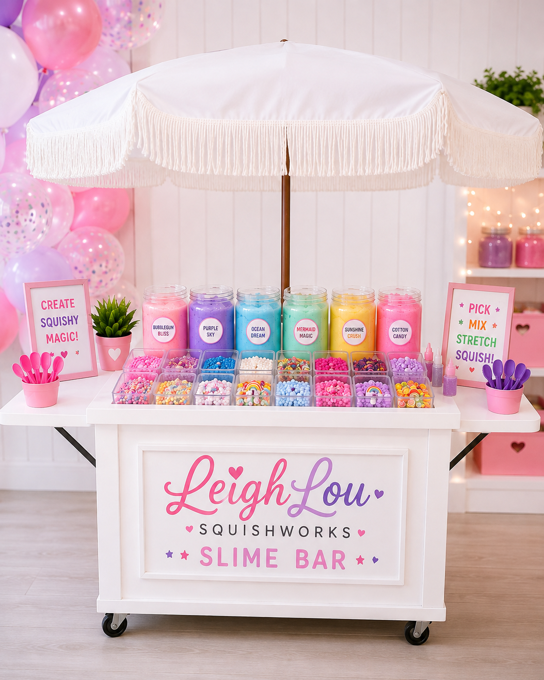 A colorful slime bar setup with containers of slime labeled with different names, sprinkles, and toppings. Signage encourages creativity and mixing. The sign on the front reads "LeighLou Squishworks SLIME BAR." There are pink and purple spoons, a small potted plant, balloons, and decorative elements in the background.