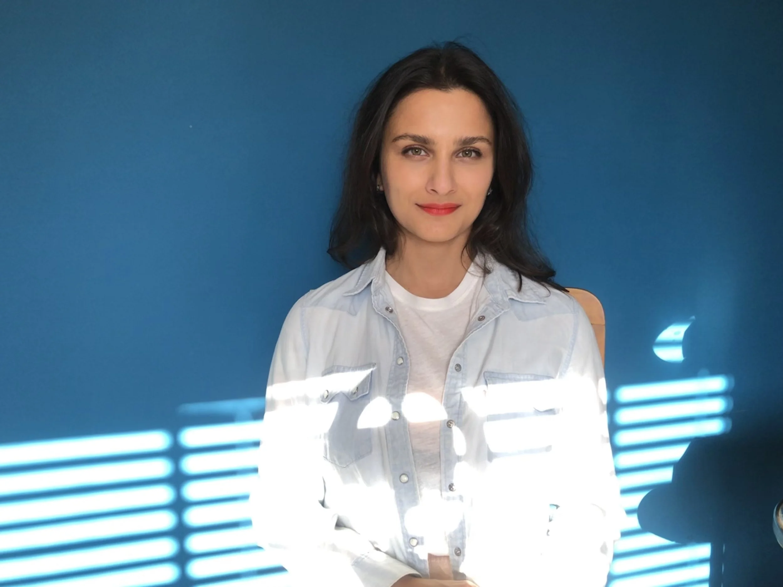 Mulher com cabelo escuro, usando camisa de botão clara, olhando para a câmera, com fundo azul e algumas luzes em primeiro plano.