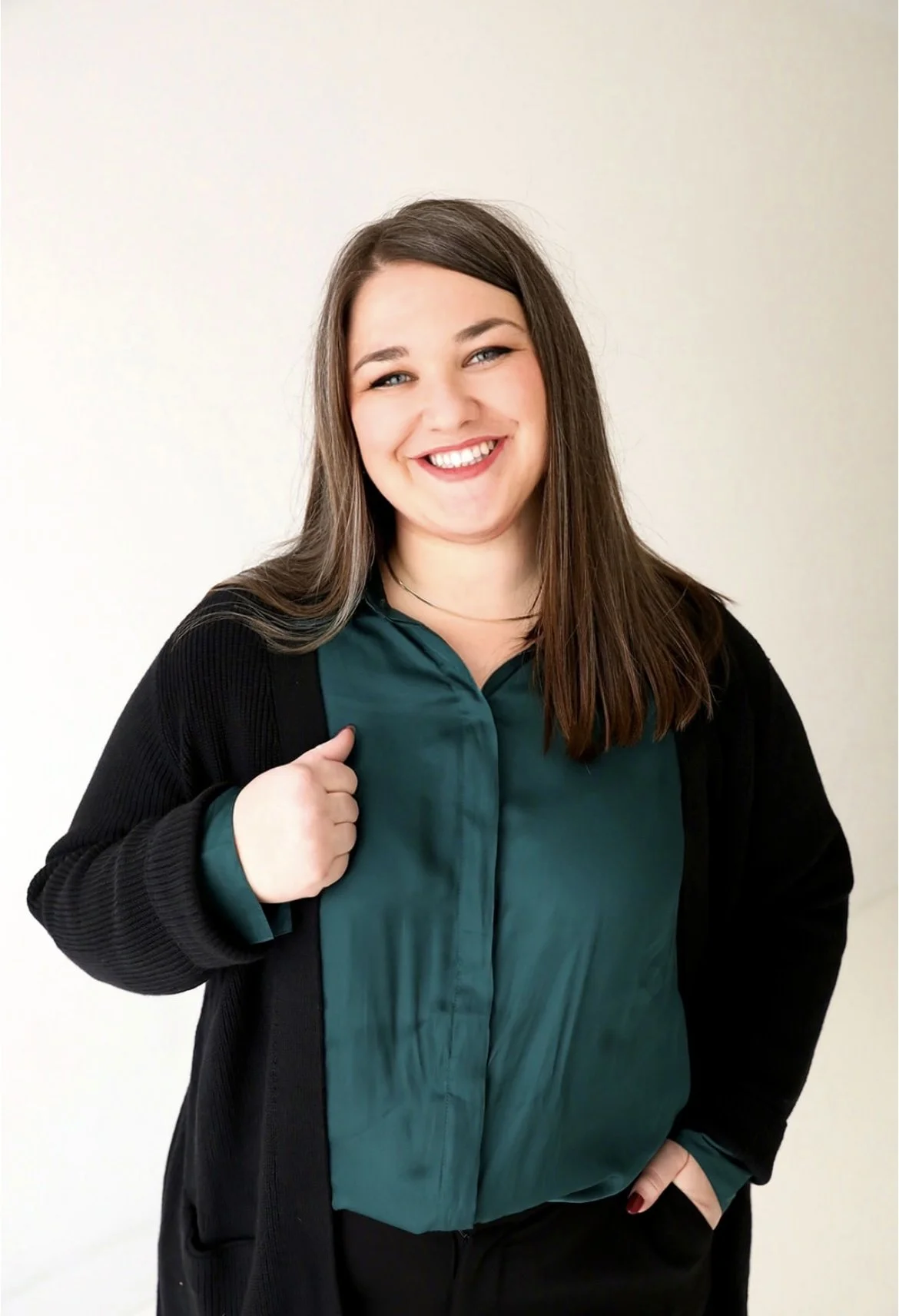 A smiling woman with brown hair, wearing a green blouse and black cardigan, standing against a plain light-colored background.