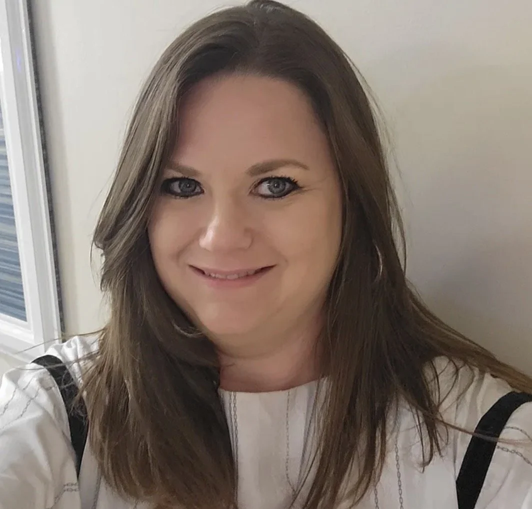 A woman with long brown hair and blue eyes taking a selfie indoors, smiling at the camera.