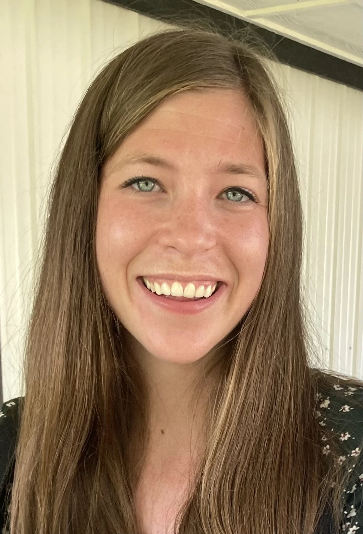 Close-up of a young woman with long brown hair, green eyes, and a big smile, standing indoors with cream-colored walls.