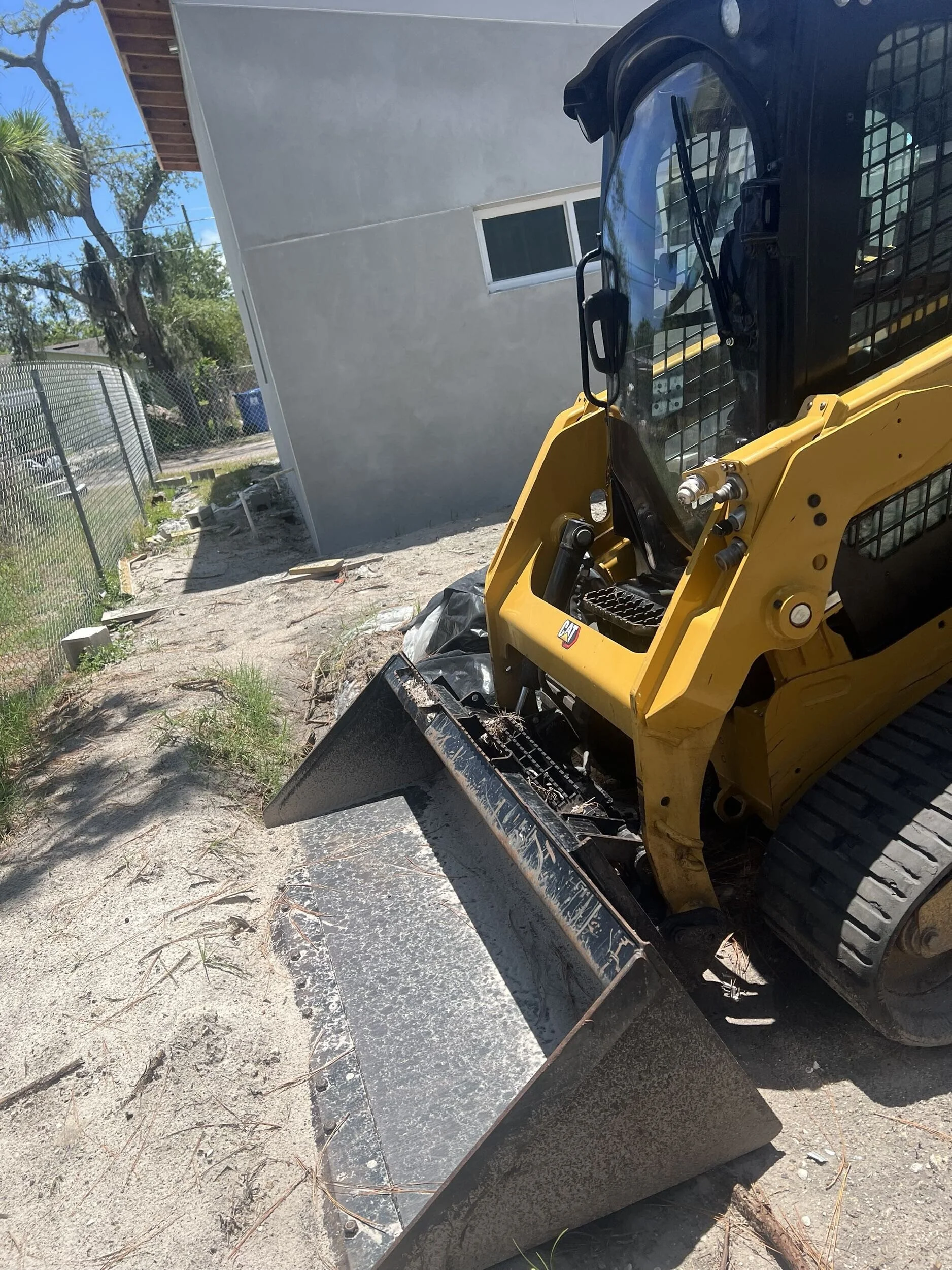 A small yellow skid-steer loader with a metal bucket attachment on a dirt and concrete surface in a backyard, next to a chain-link fence and gray house wall, with trees and a blue sky in the background.