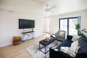 Living room with white walls, a large TV on the wall, a dark sofa, a smaller matching sofa, a black coffee table, a ceiling fan, a sliding glass door, and a potted plant.