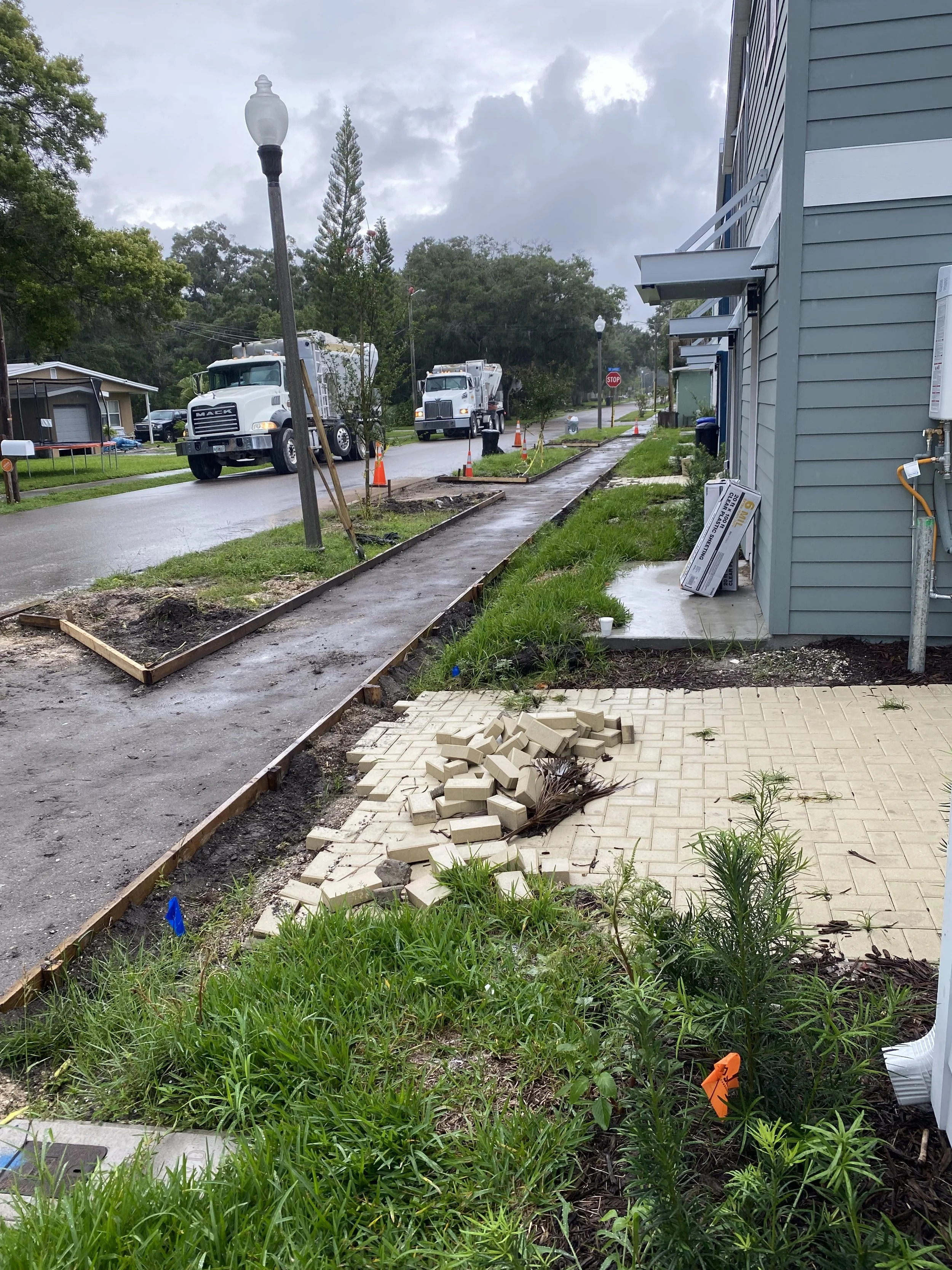 A residential street under construction with a new sidewalk, orange cones, and two white trucks. There is a green lawn with plants and a small tree to the right, and a blue house on the right side of the street. The sky is cloudy and overcast.