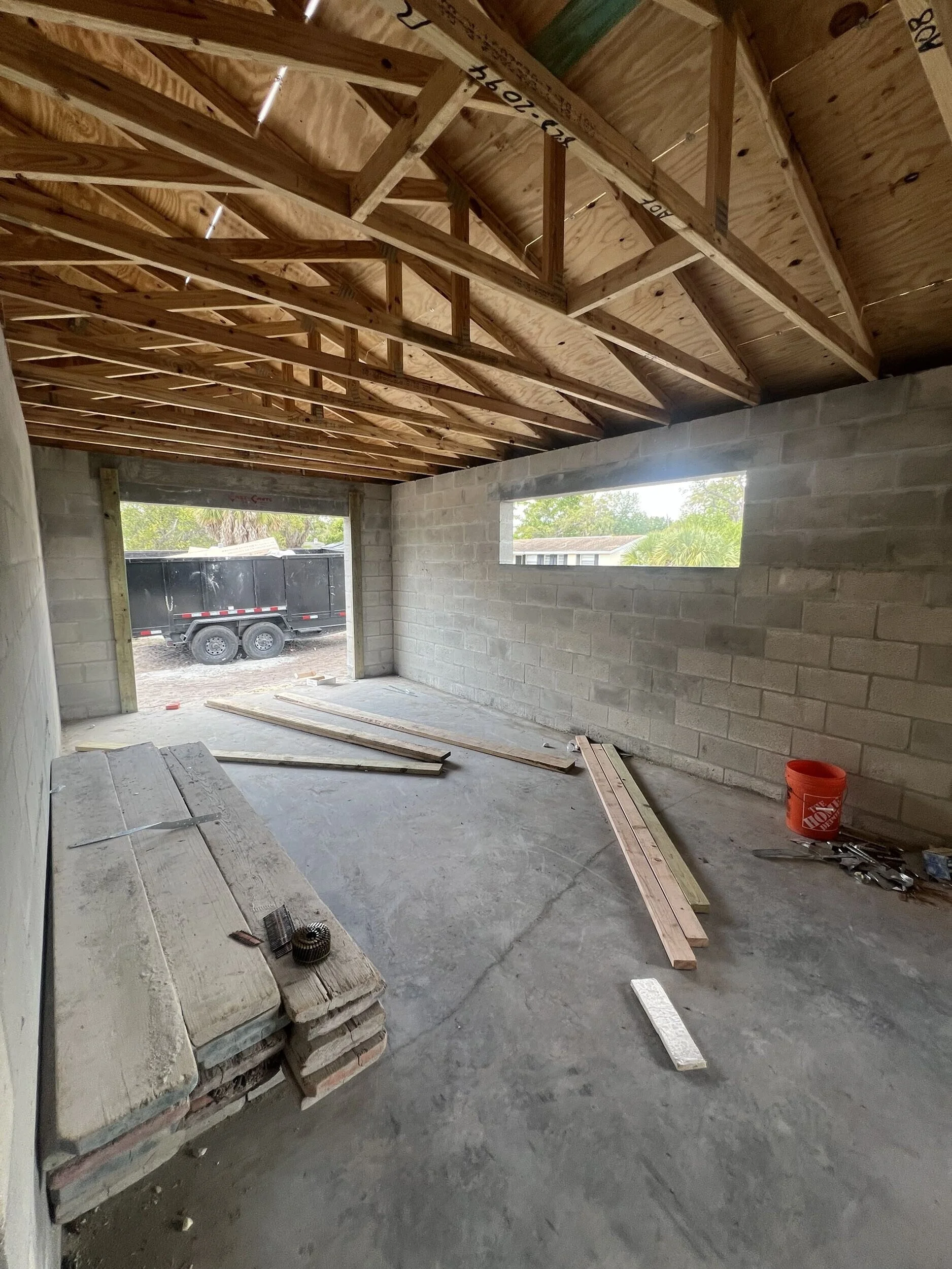 Interior of a house under construction with exposed wooden roof trusses, unfinished concrete block walls, construction tools, wood planks, and a black truck visible outside through an open doorway.