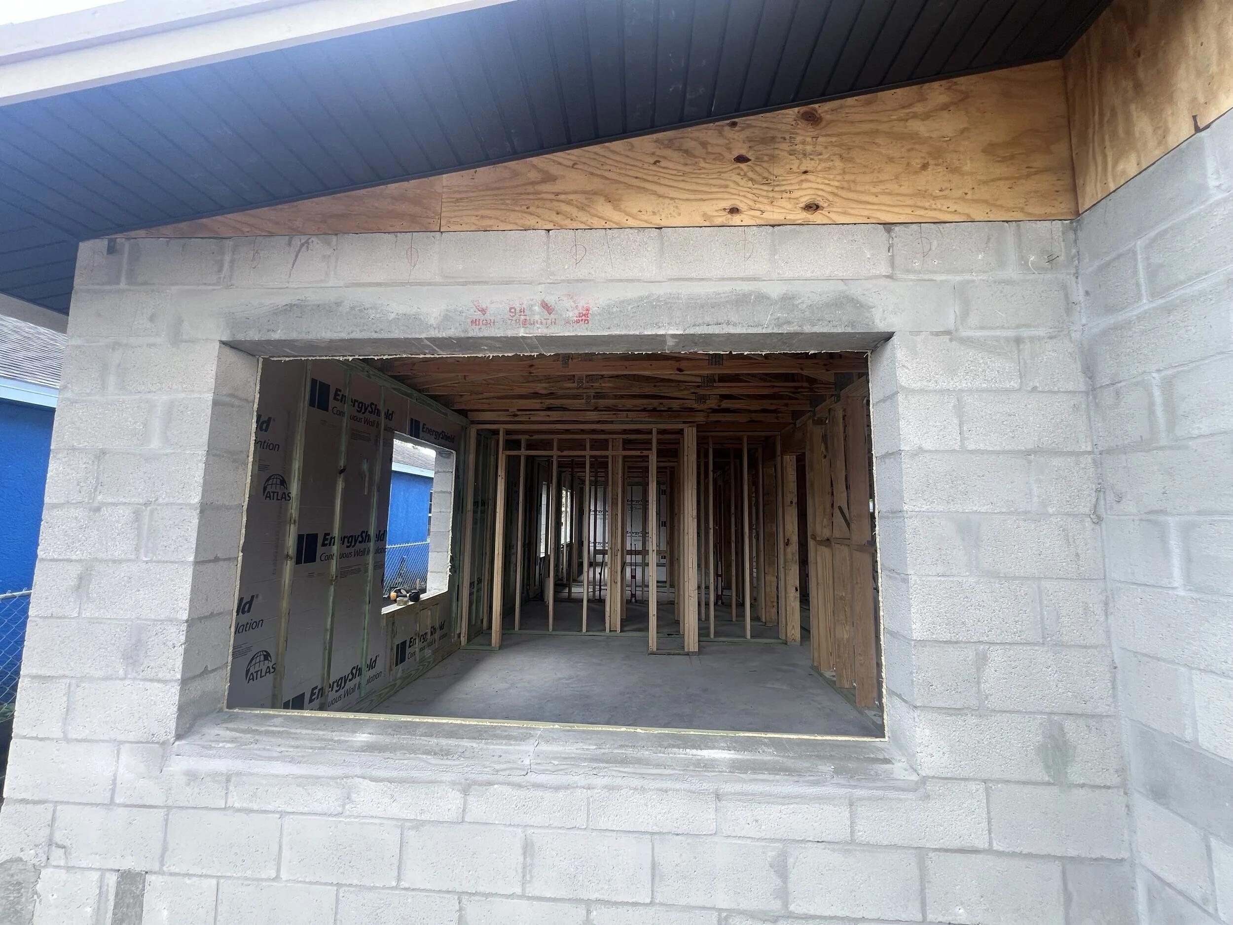Construction site showing window opening framed in concrete block wall, interior with wooden framing and roof look-up.