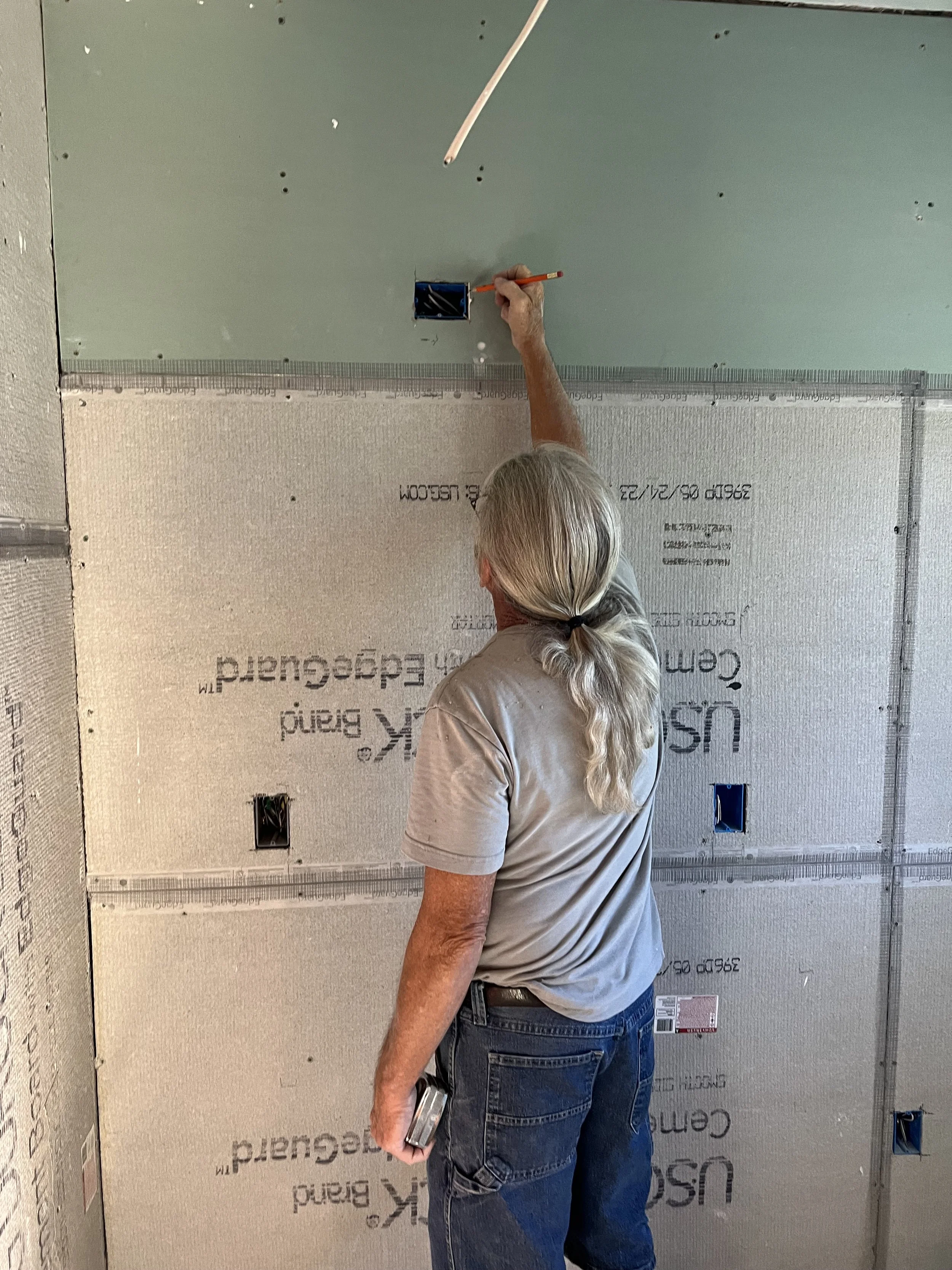 A person with gray hair in a ponytail installing electrical wiring in a wall with drywall. The person is facing the wall and holding a tool while reaching into a small electrical box on the wall.