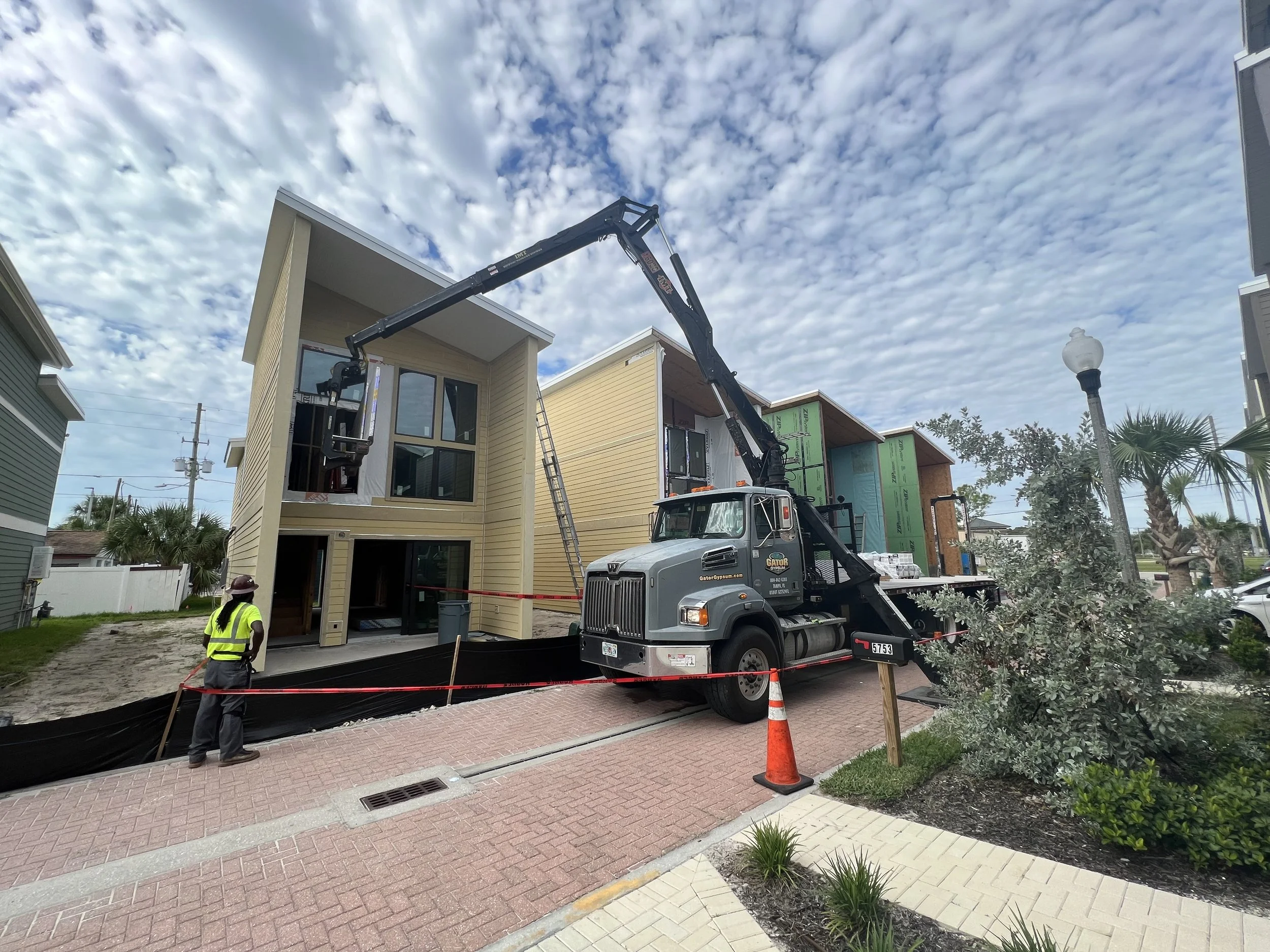 A construction site with a crane truck lifting a house section, two workers, and a partly constructed building, under a partly cloudy sky.