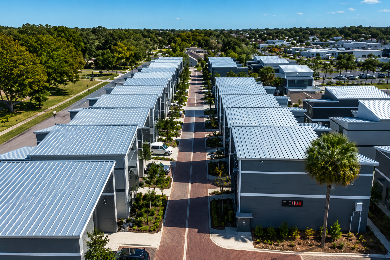 Aerial view of a modern residential complex with two parallel rows of townhouses with metal roofs, a brick-paved central walkway, parked cars, landscaped trees, and a park with large trees in the background.
