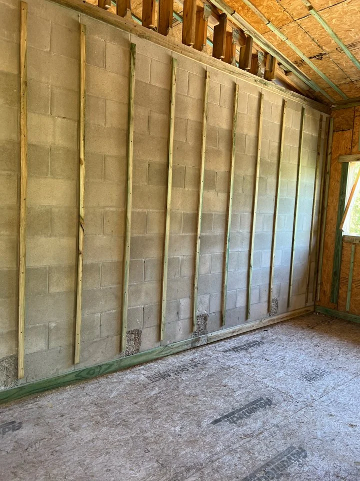 Interior of a building under construction with a partially framed wall with vertical wooden studs, cinder block wall, and exposed ceiling framing.