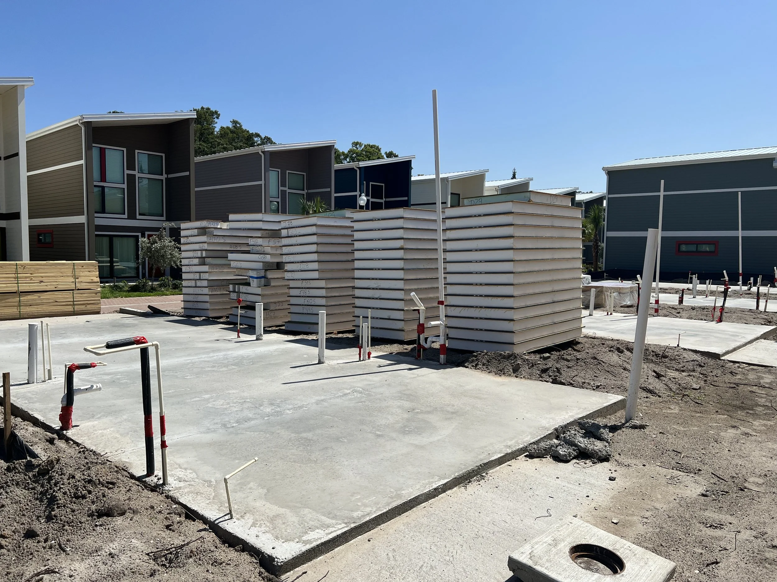 Construction site with multi-story modern residential buildings, stacks of insulation panels, wooden planks, and construction materials, under a clear blue sky.