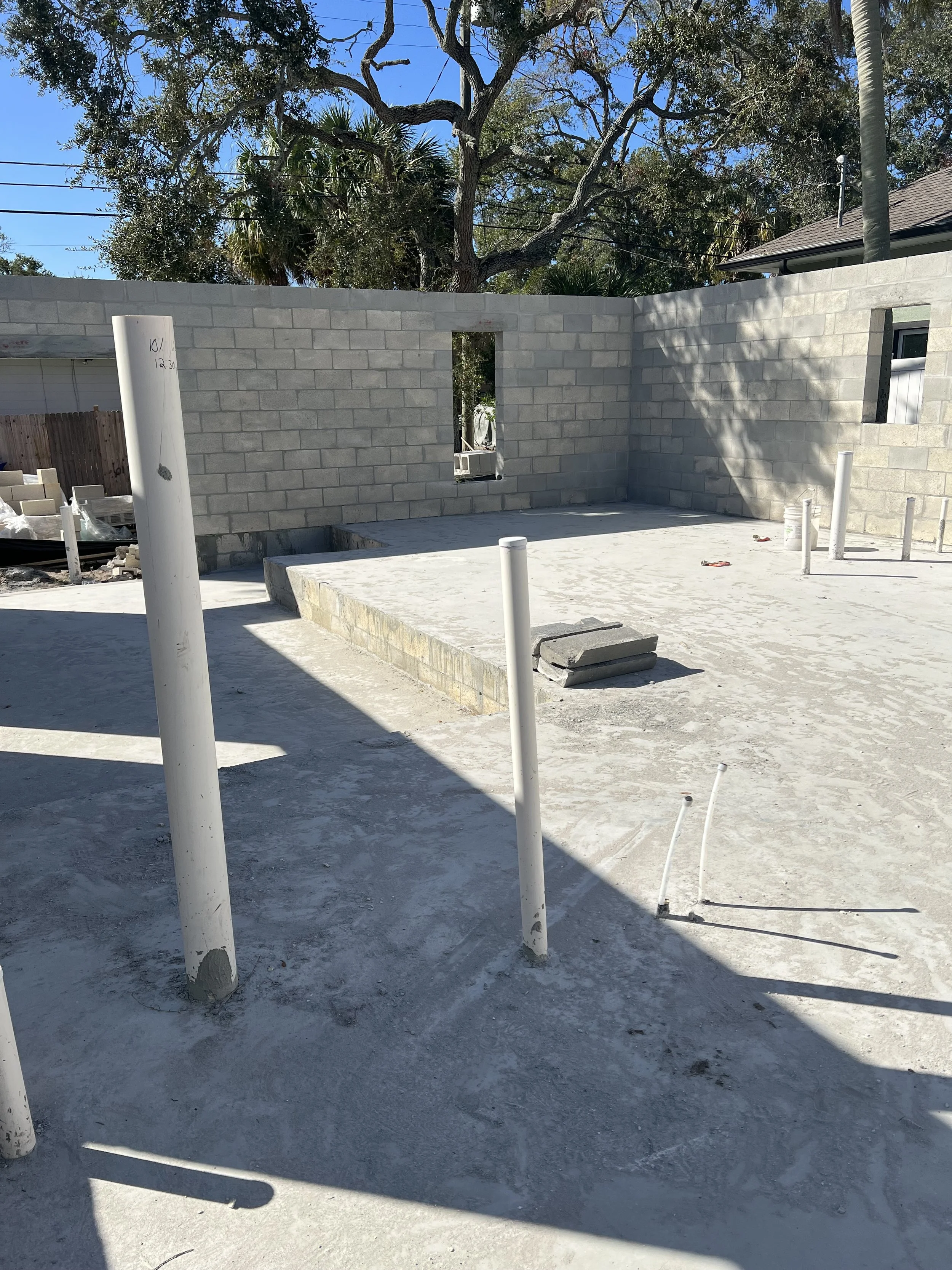 Construction site with unfinished concrete block walls, exposed pipes, and scattered construction materials, under a clear blue sky with trees in the background.