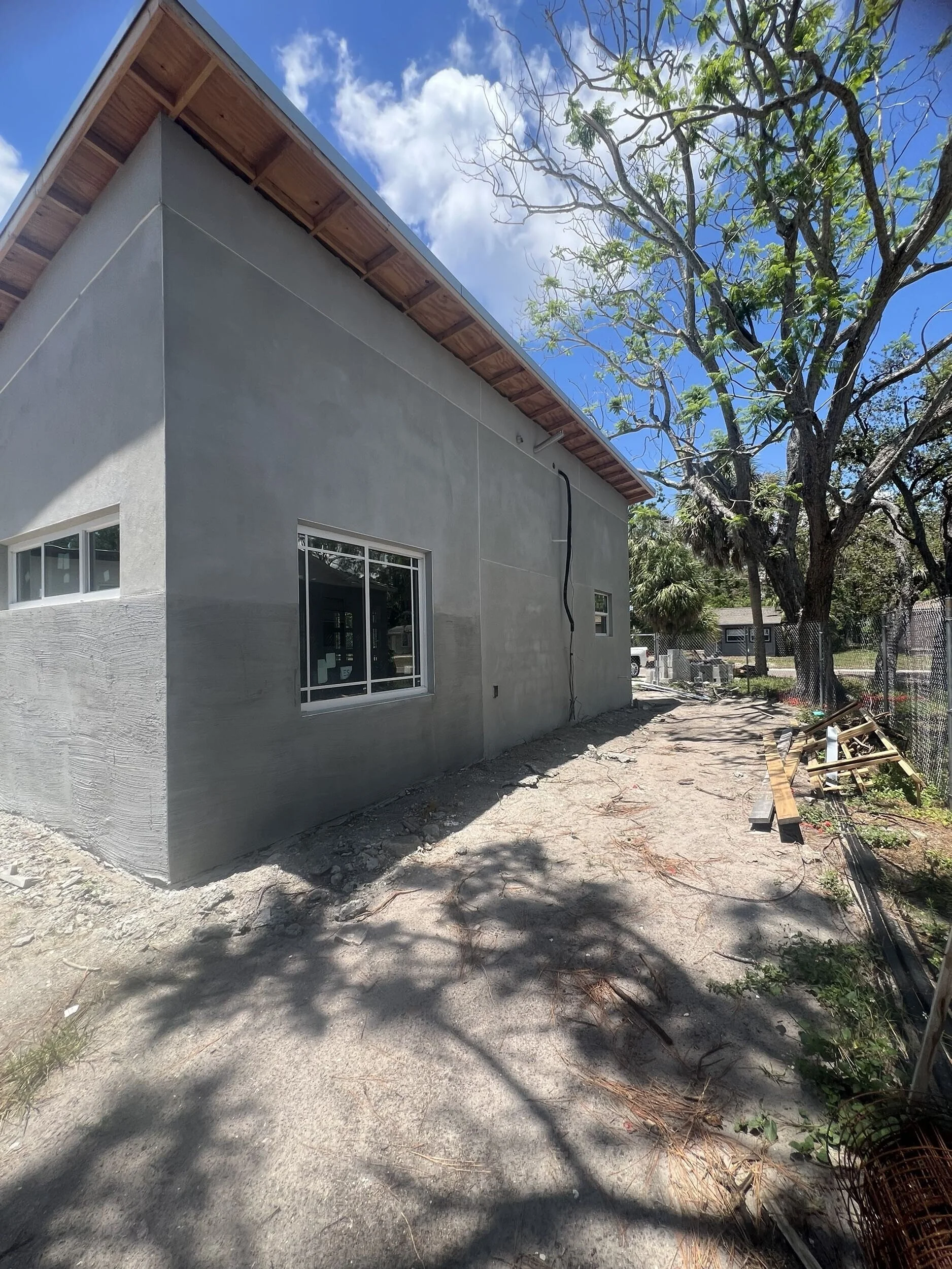 Side view of a house under construction with a newly finished exterior wall and window, construction debris, and a large tree with green leaves and some bare branches nearby, under a partly cloudy sky.