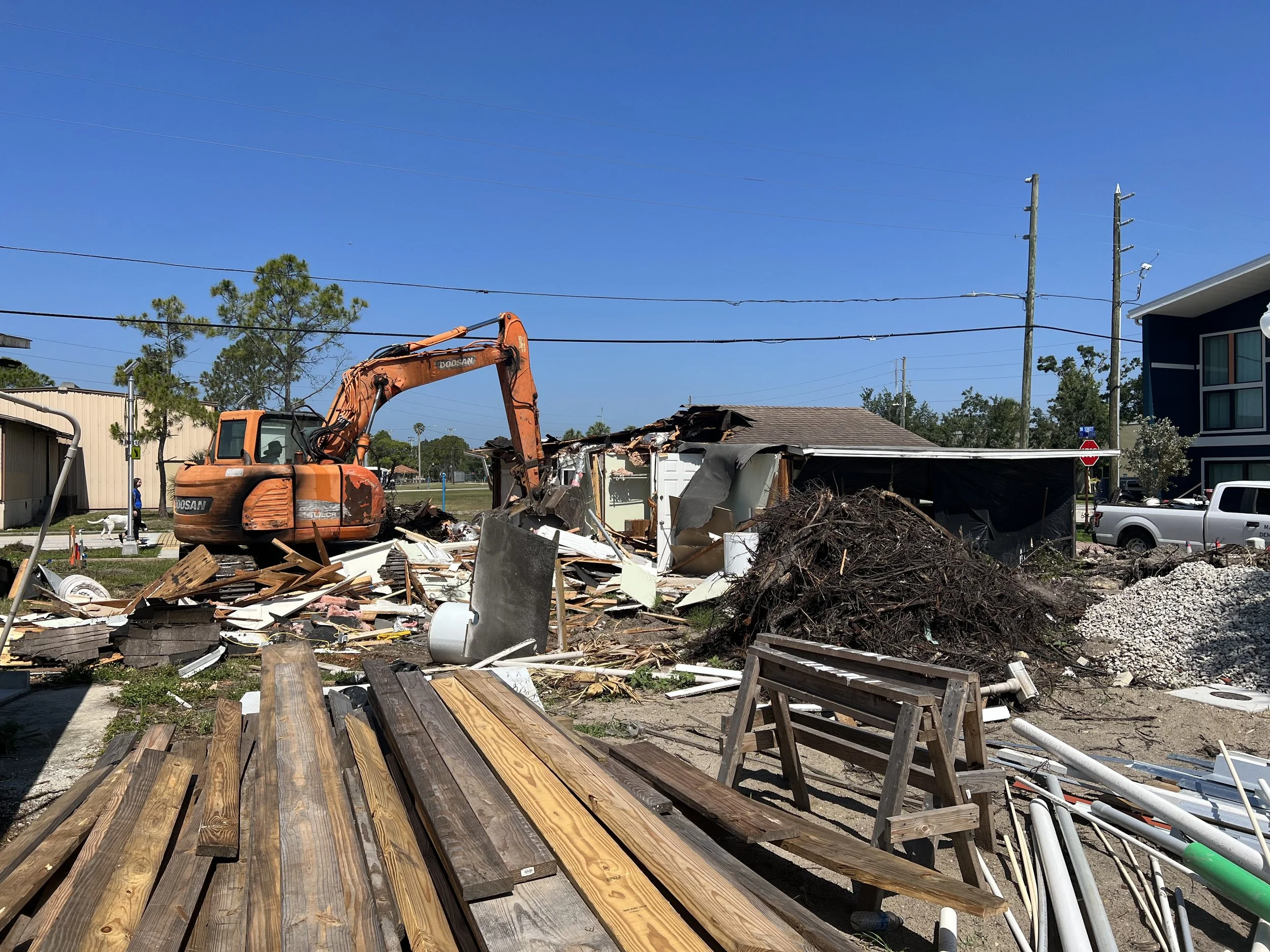 A construction or demolition site with an orange excavator, debris, and a partially demolished building. Wooden planks are in the foreground, and power lines and trees are in the background under a clear blue sky.