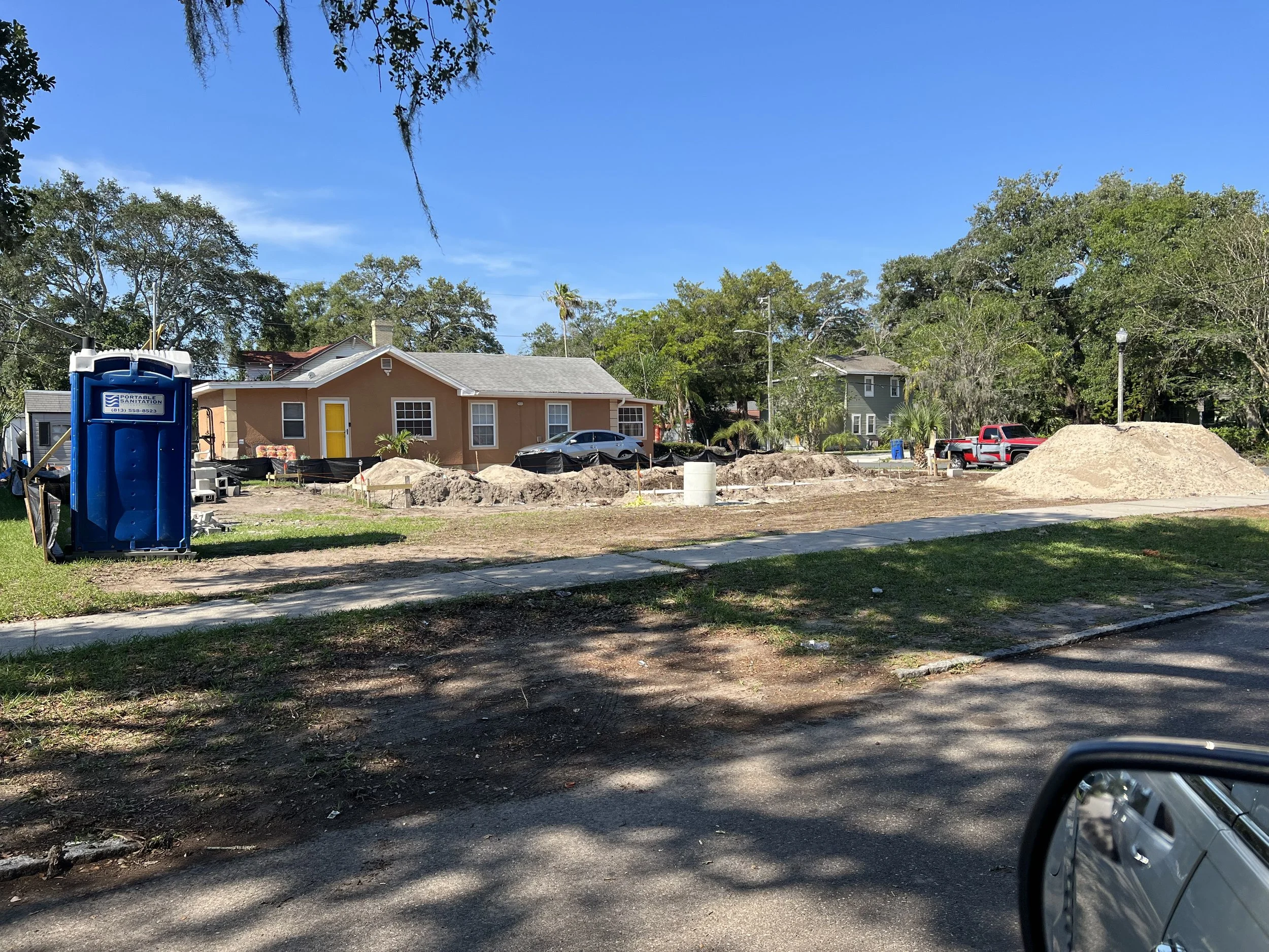 A construction site in a suburban neighborhood with piles of dirt, a portable sanitation unit, and a partly constructed house with a yellow door. There are trees, parked cars, and a sidewalk in the background with a clear blue sky.