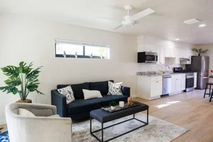 Living room with a dark blue sofa, white armchair, coffee table, and kitchen in the background.