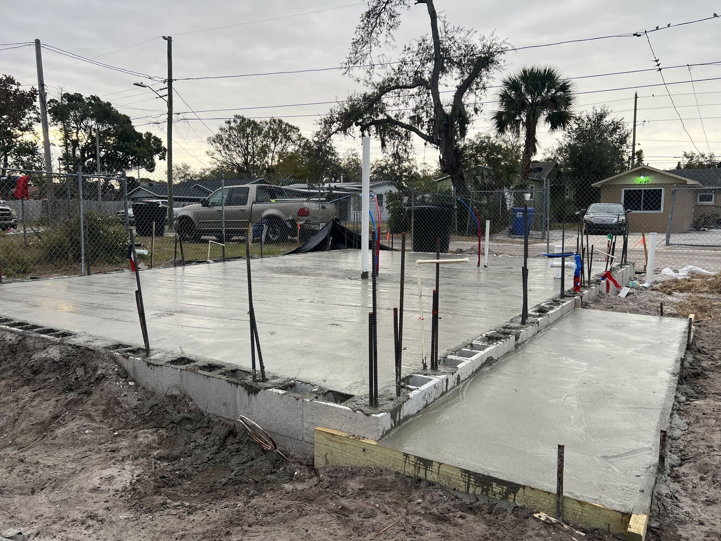 Concrete foundation slab with rebar and wooden form boards, construction site, residential neighborhood with trees and power lines in the background.