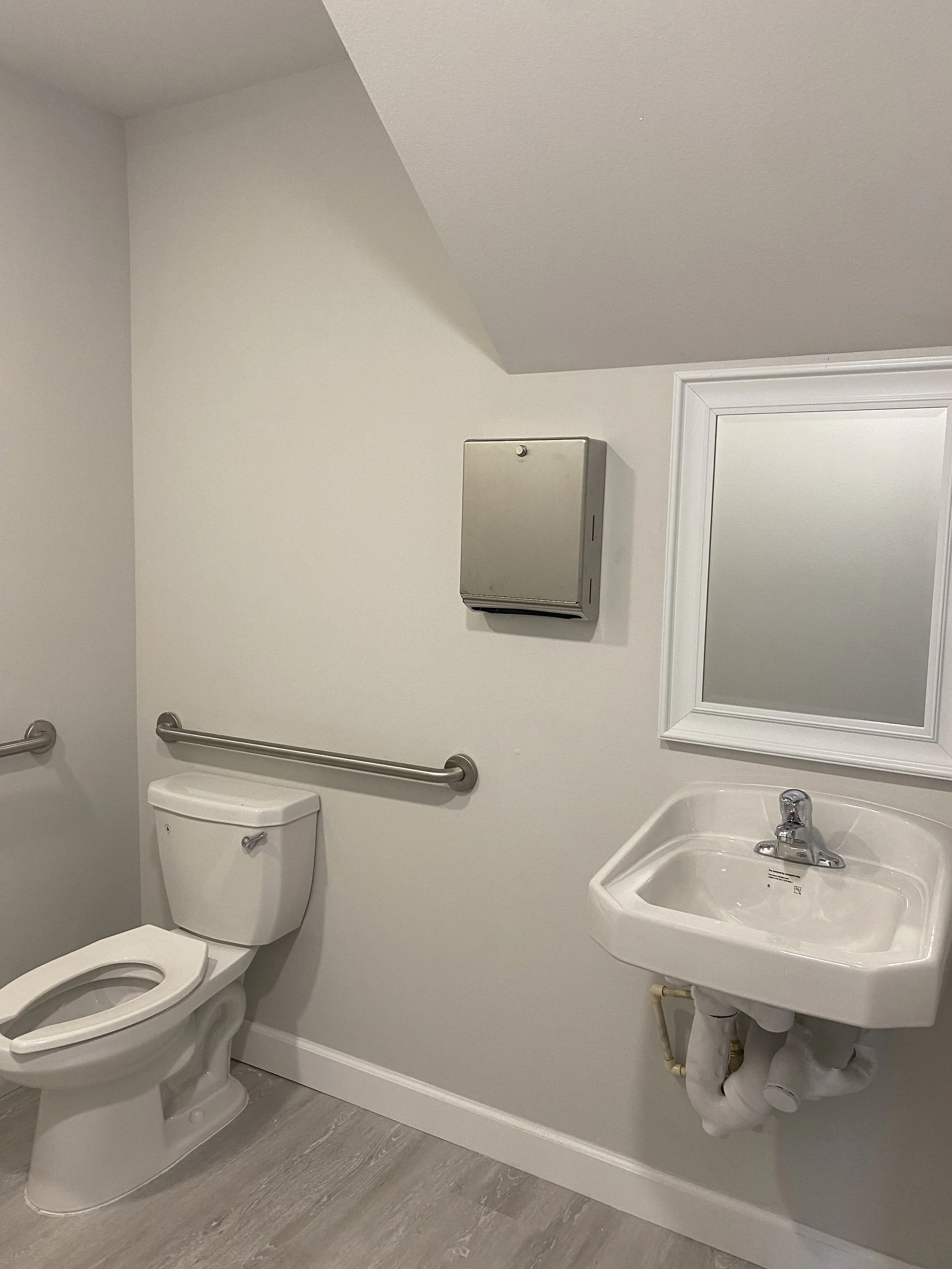 A bathroom with a white toilet, a wall-mounted stainless steel hand dryer, a mirror with a white frame, a small sink with a single faucet, and grab bars on the wall. The floor has light gray wood-look flooring.