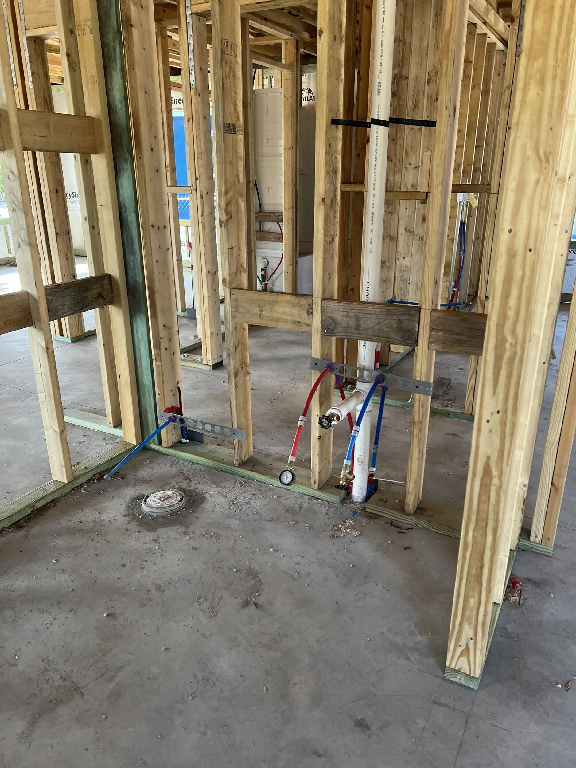 Interior wooden framing of a building under construction, with visible plumbing pipes, electrical wiring, and a floor drain.