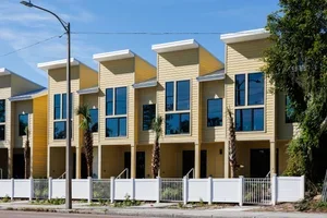 Row of modern townhouses with yellow and beige exteriors, large blue windows, and white fencing.