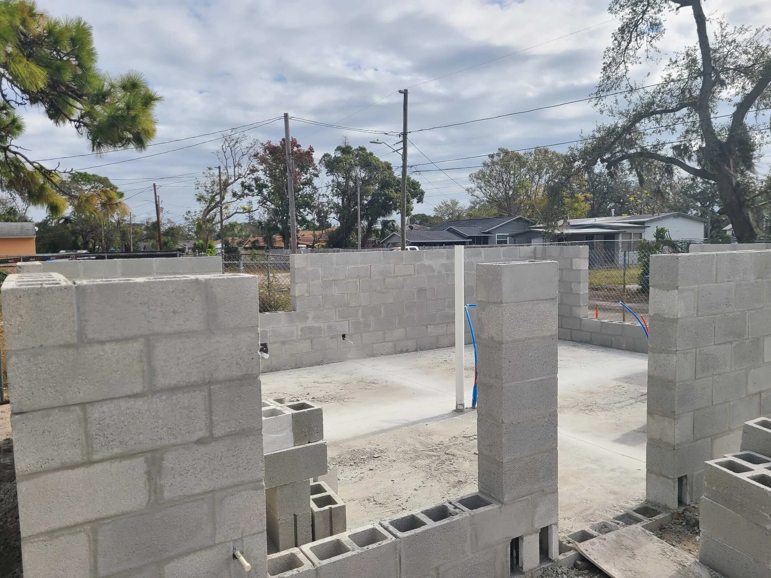 Construction site with unfinished concrete block walls, concrete foundation, electrical wiring, and nearby residential houses under a cloudy sky.
