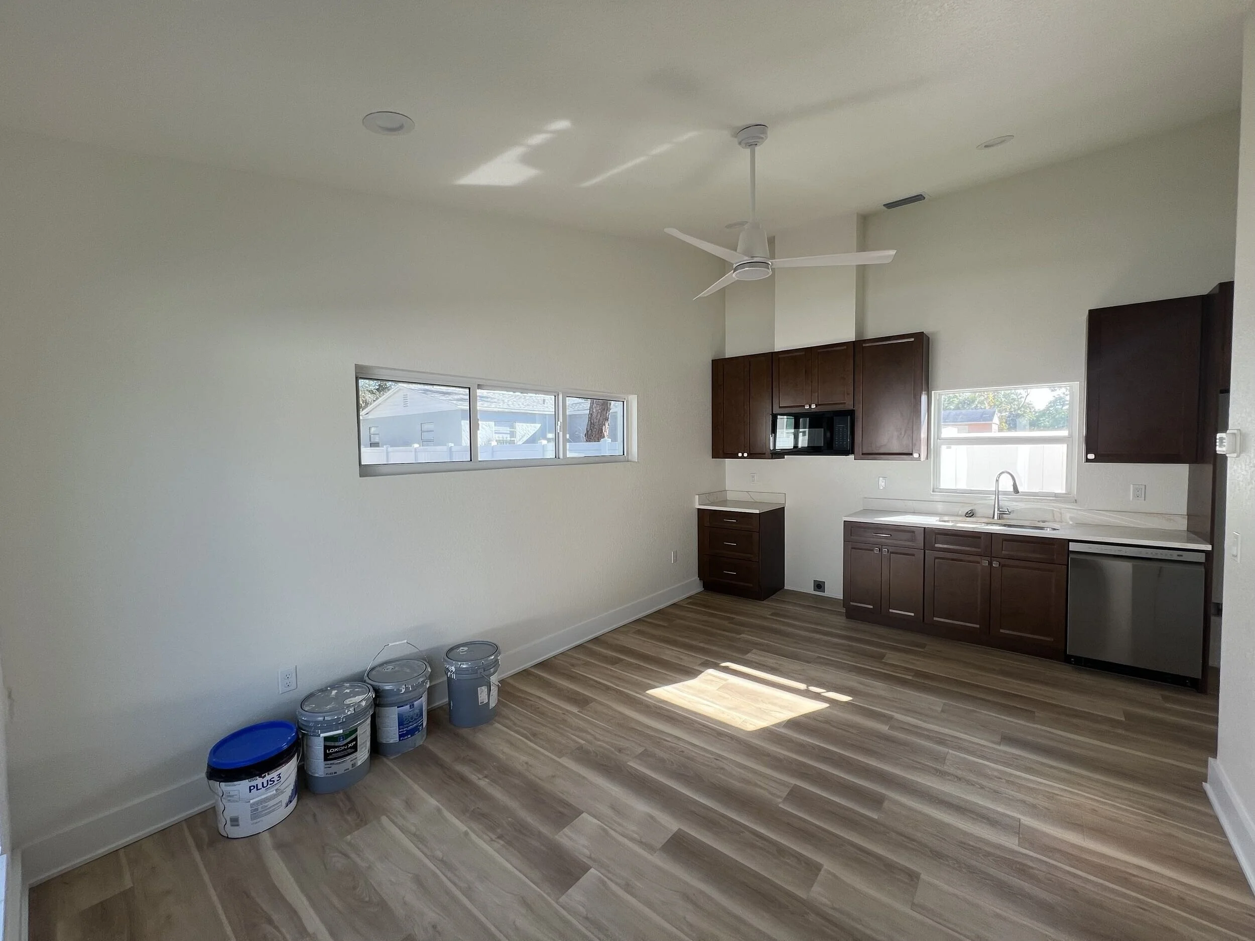 A room with a ceiling fan, dark wooden cabinets, a window showing a house and trees outside, and several buckets of paint on the floor.