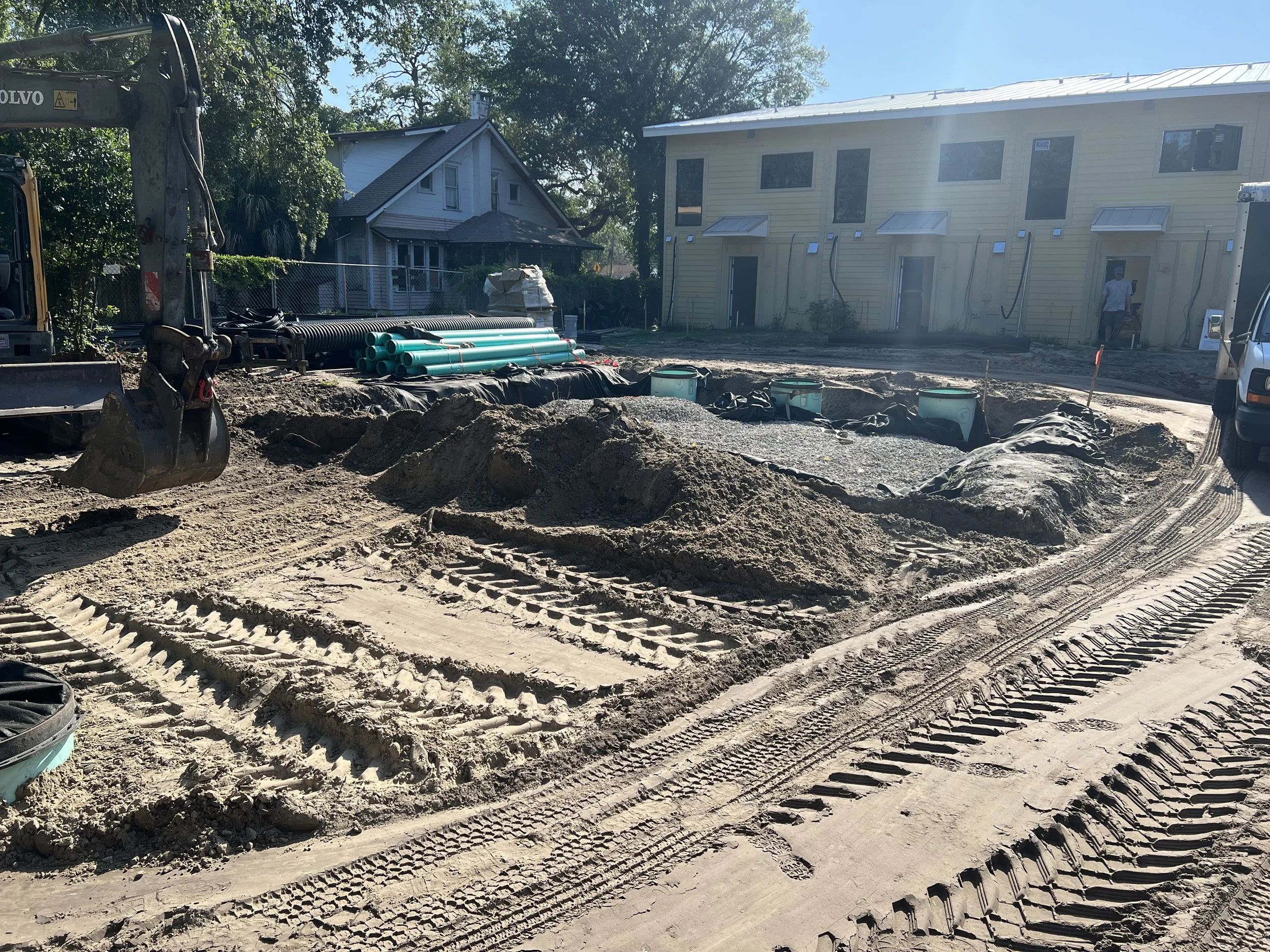 Construction site for a residential building with dirt, construction equipment, pipes, and two houses in the background.