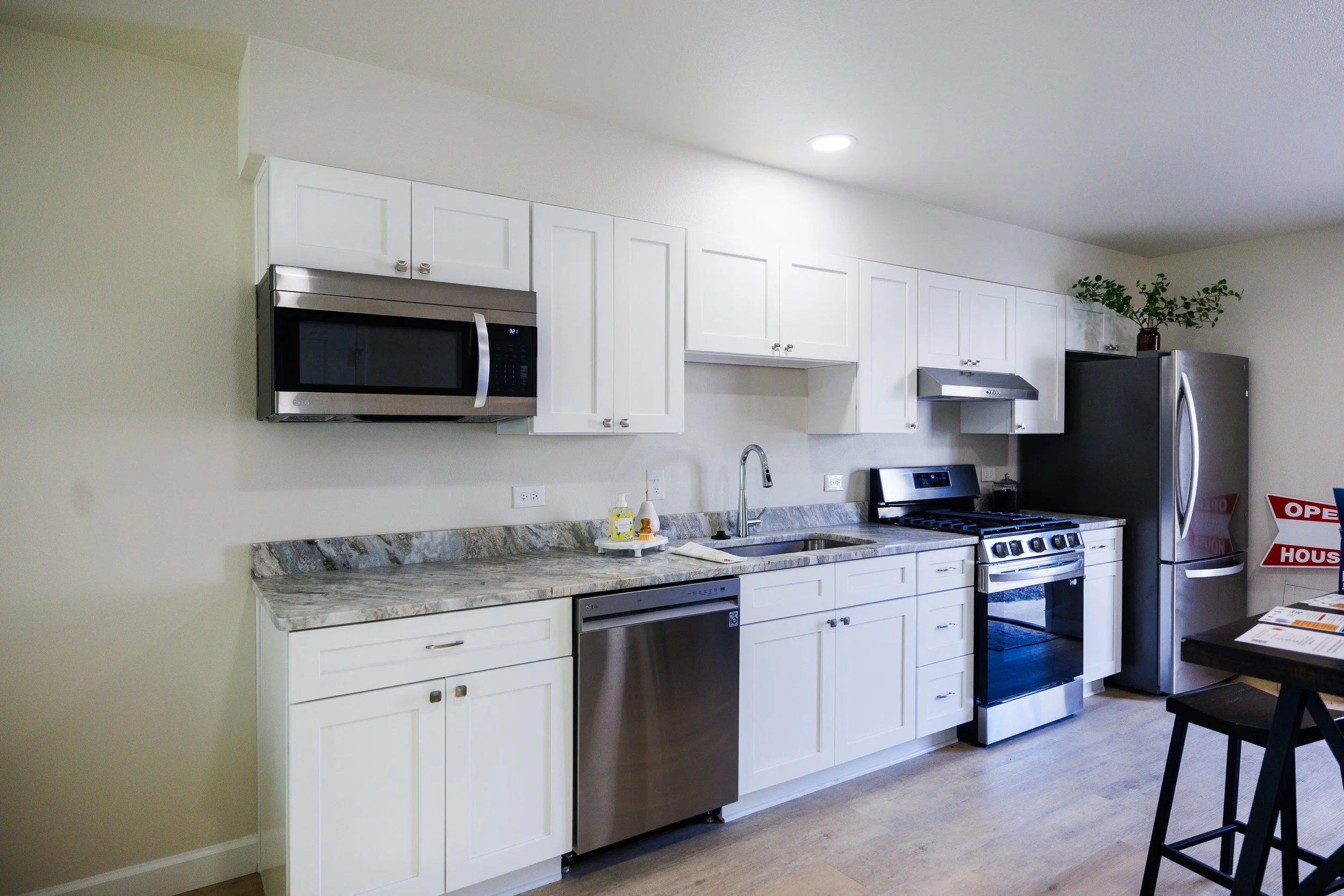 Kitchen with white cabinets, stainless steel microwave, range, refrigerator, and a marble countertop. There is a potted plant on top of the cabinets and a black chair at the dining table.