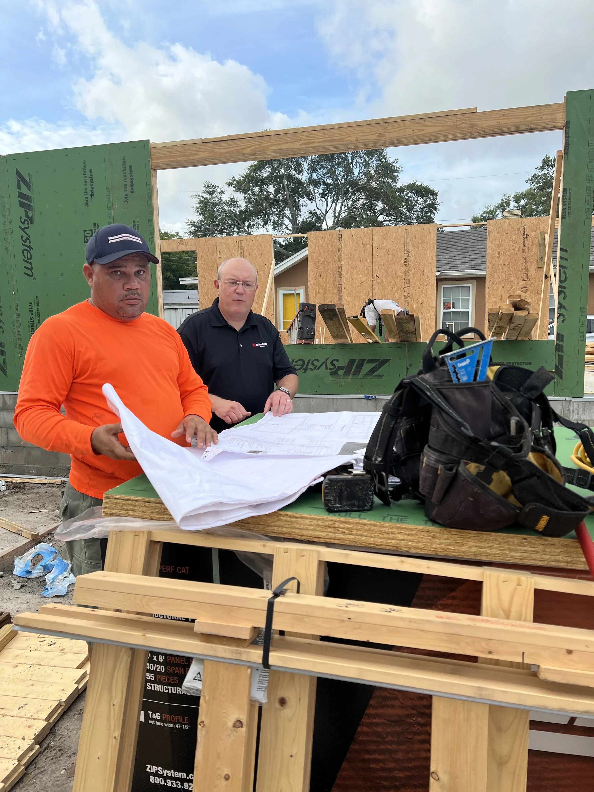 Two men are standing in front of a partially constructed building. One man is wearing an orange shirt and a baseball cap, holding construction plans. The other man is wearing a black shirt, also with plans on the table. There is construction equipment and lumber on a work table in front of them, and a partially built wall with green sheathing behind them.