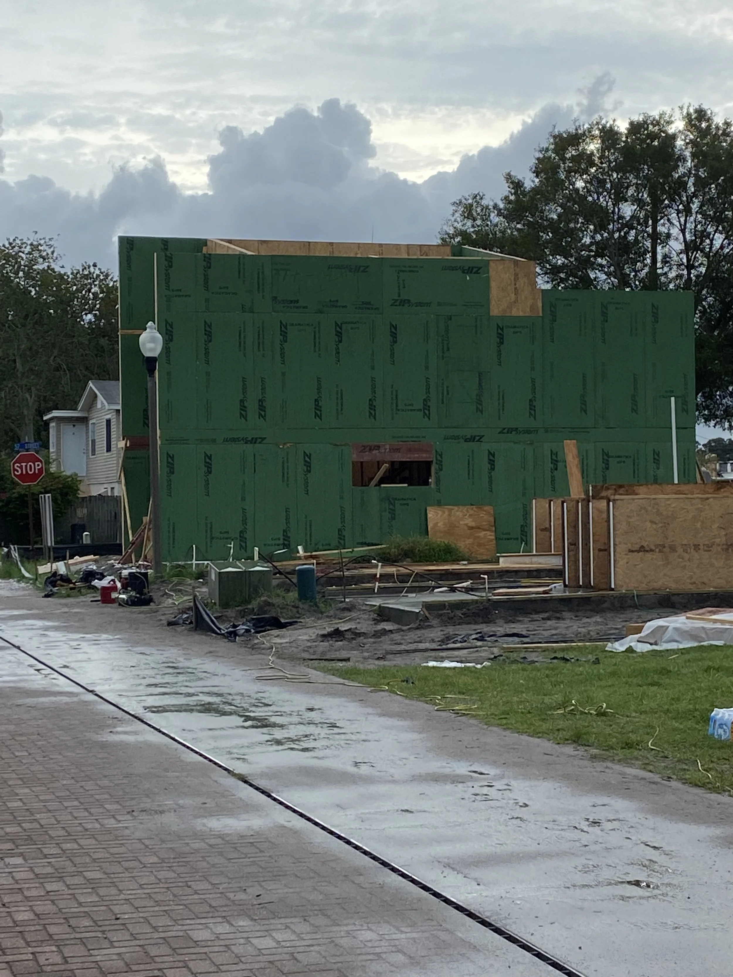 A house under construction with green sheathing on the walls, scattered construction materials and tools on the ground, and a cloudy sky overhead.
