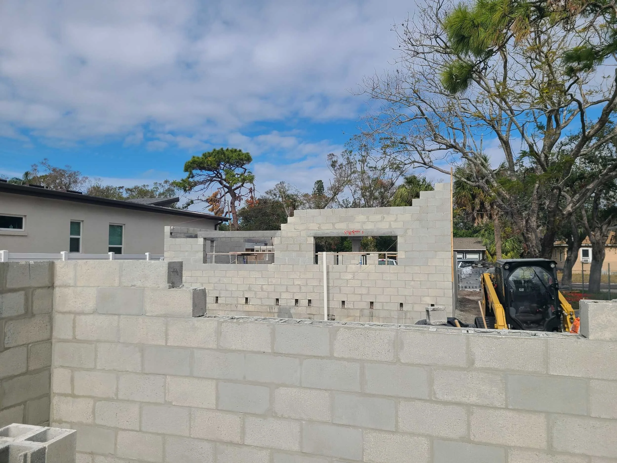 Construction site with a partially built brick wall, surrounding trees, and a building in the background under a cloudy sky.