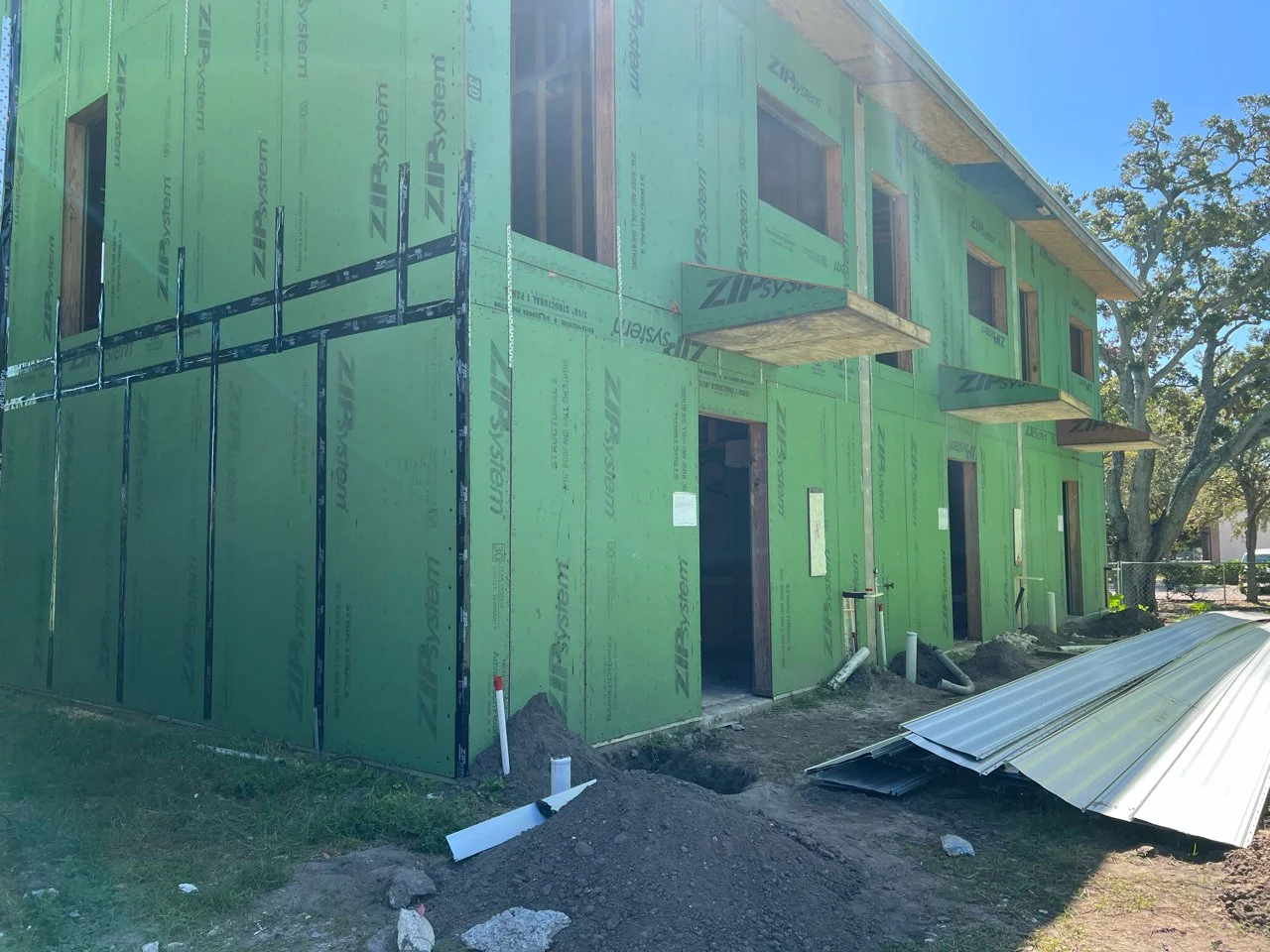 Green construction sheathing on a building under construction, with open window frames, building materials on the ground, and blue sky overhead.