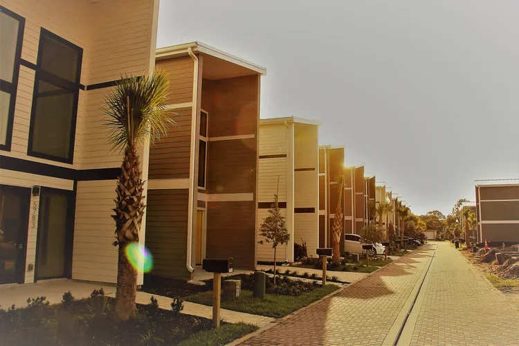 A row of modern townhouses with balconies, palm trees, and a paved sidewalk on a sunny day.
