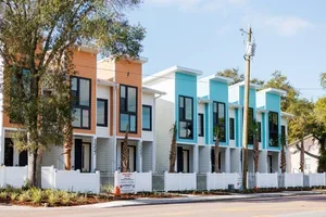 Row of modern townhouse buildings with colorful facades, trees, and a sidewalk in the foreground.