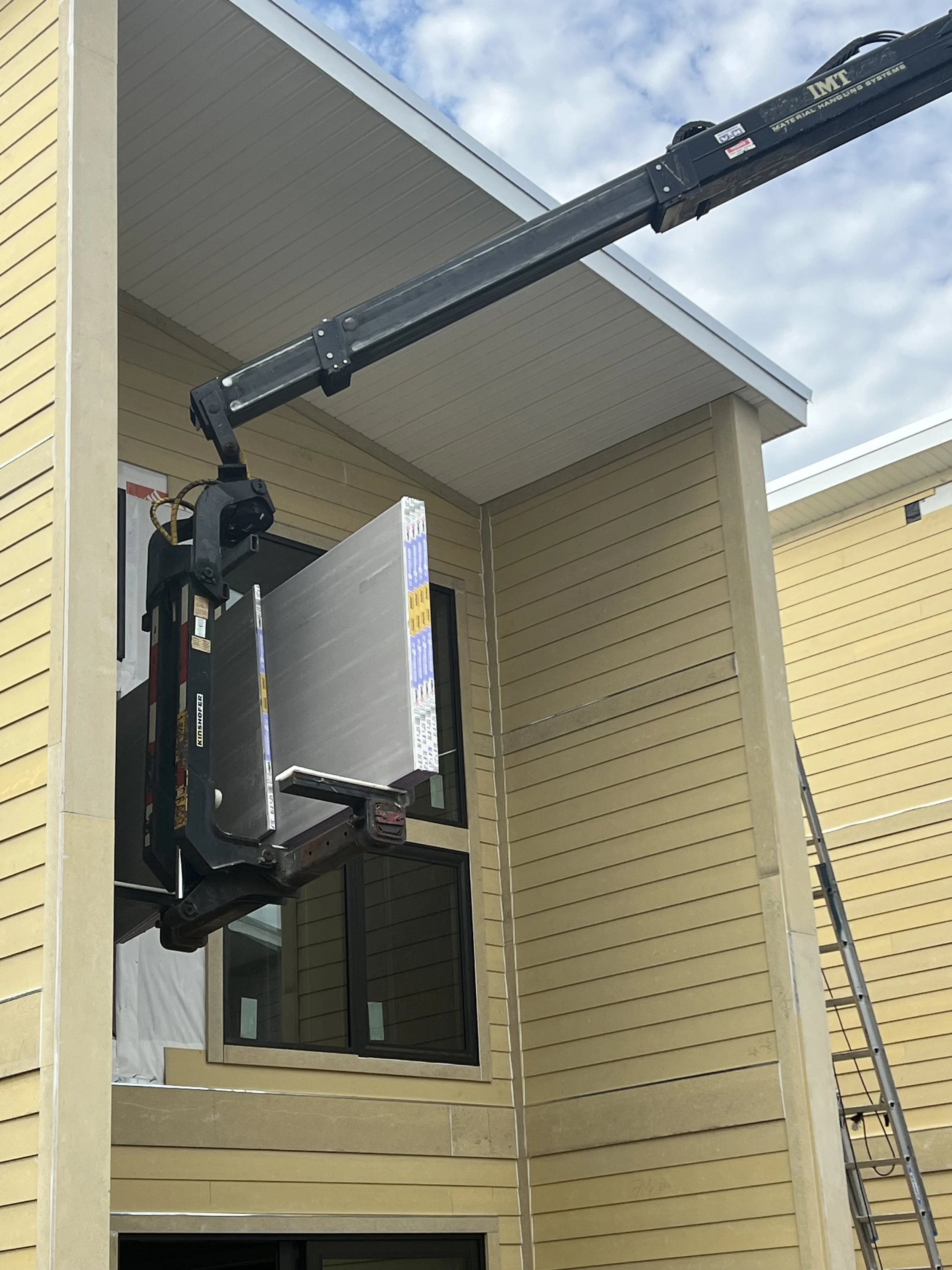 Construction worker installing a new window in a yellow house using a hydraulic lift, with a blue sky and clouds in the background.