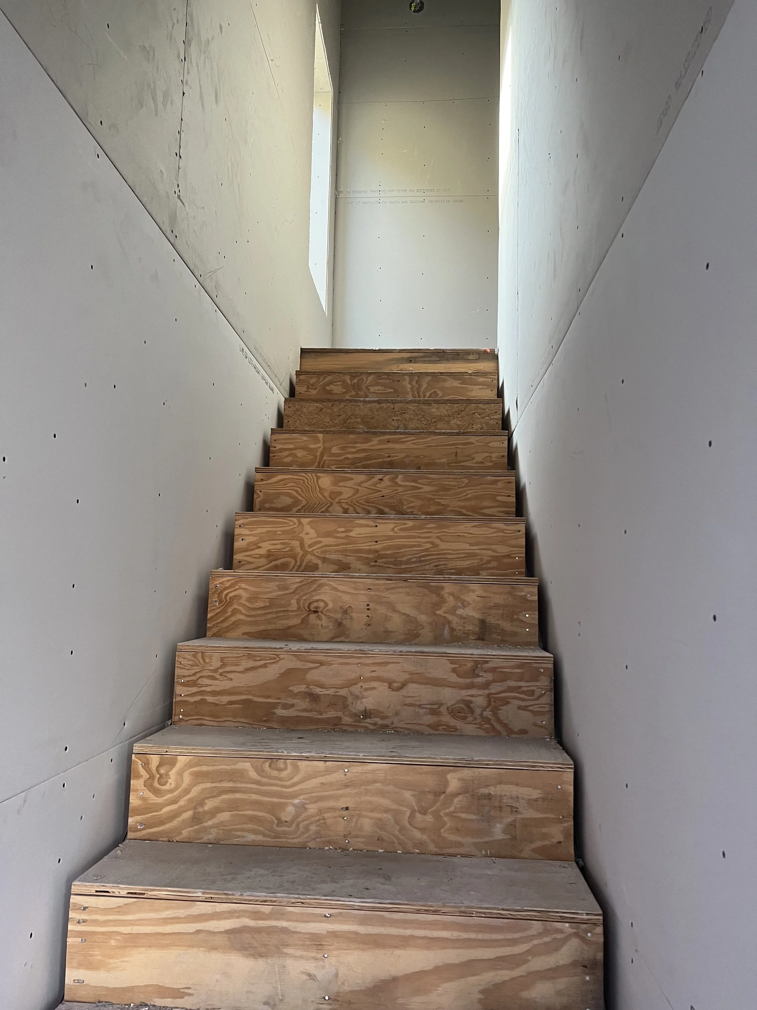 Wooden staircase with unfinished plywood steps in a narrow, partially built hallway.