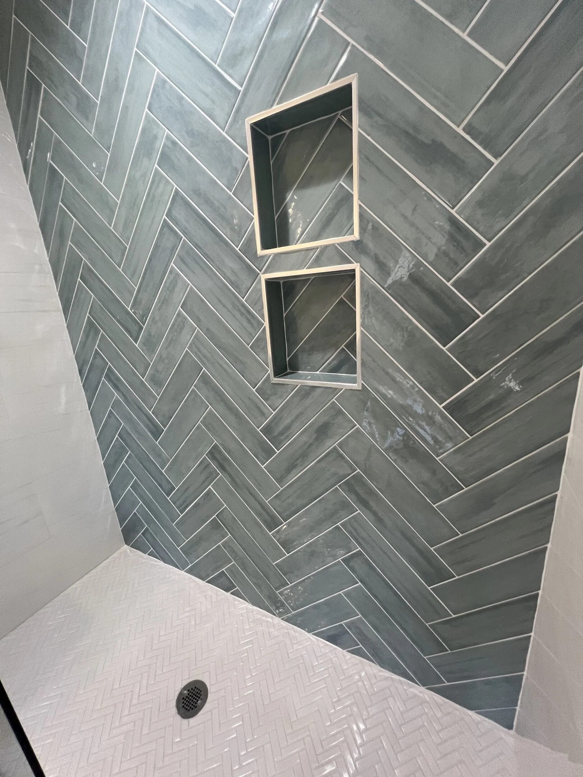 Shower area with dark gray herringbone tile walls, a white herringbone tile floor, a drain, and two built-in rectangular shelves in the wall.