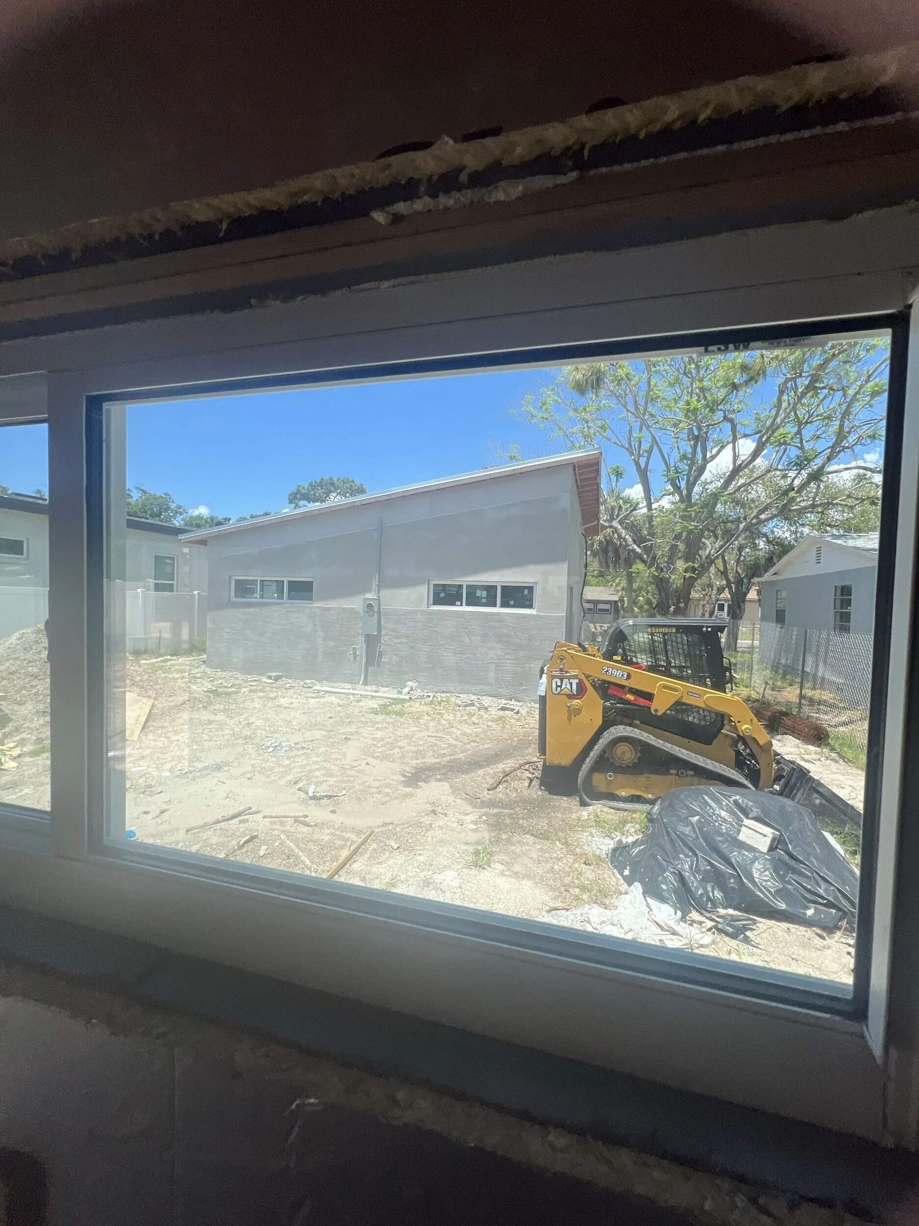 Residential backyard with construction equipment, trees, and nearby houses, viewed through a window.