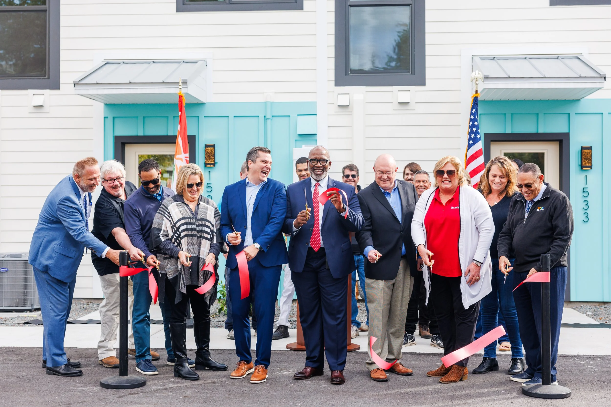 A group of diverse people at a ribbon-cutting ceremony outside a modern house with white siding and blue accents, with American flags.