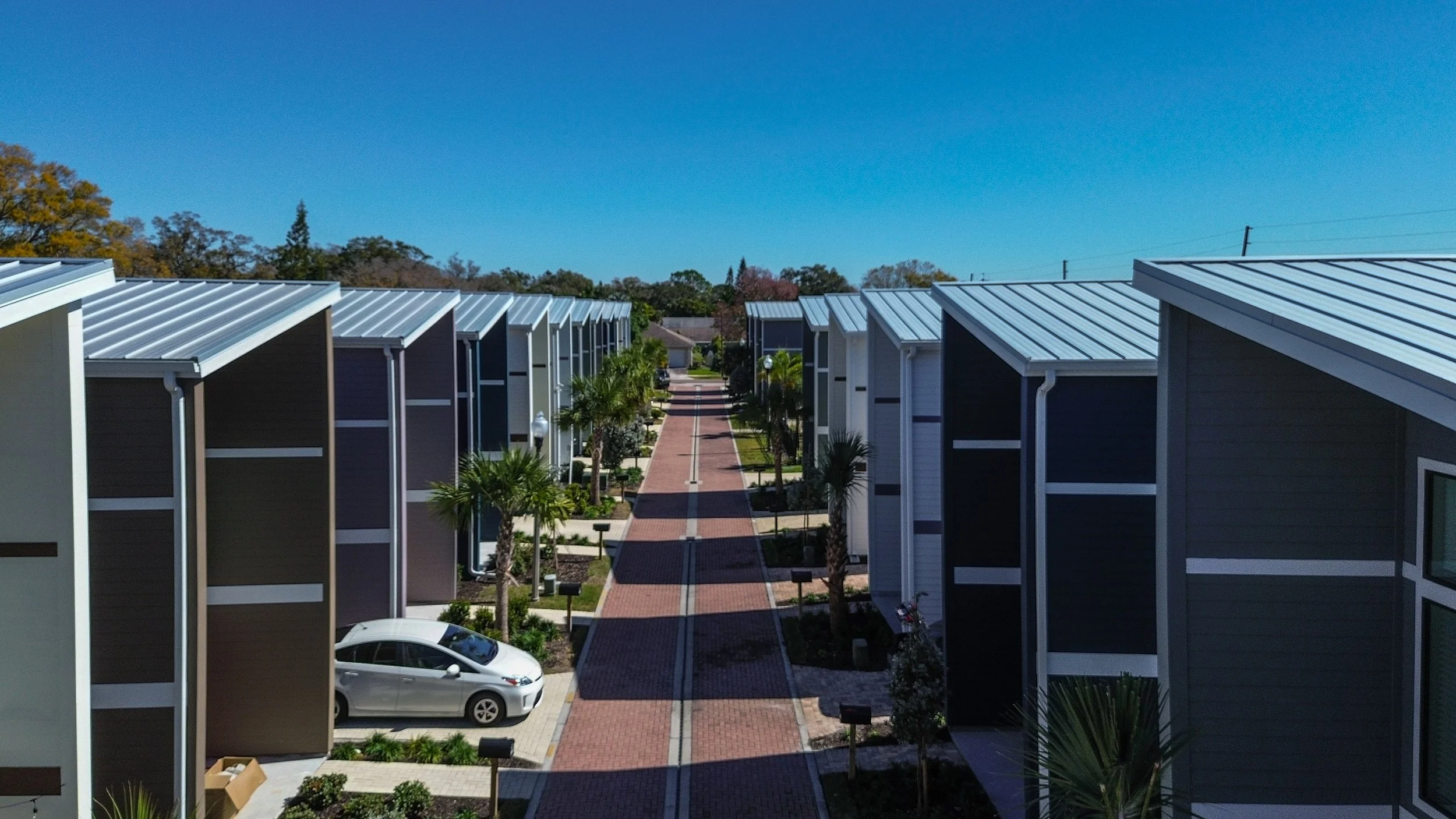 A residential neighborhood with modern townhouses featuring metal roofs, a brick-paved walkway, small palm trees, and a parked silver car in the foreground.