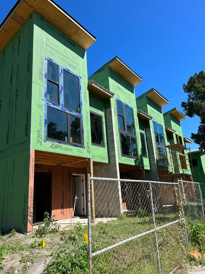 Construction site with multi-story buildings in progress, green sheathing on exterior walls, large windows, and scaffolding, under a clear blue sky.