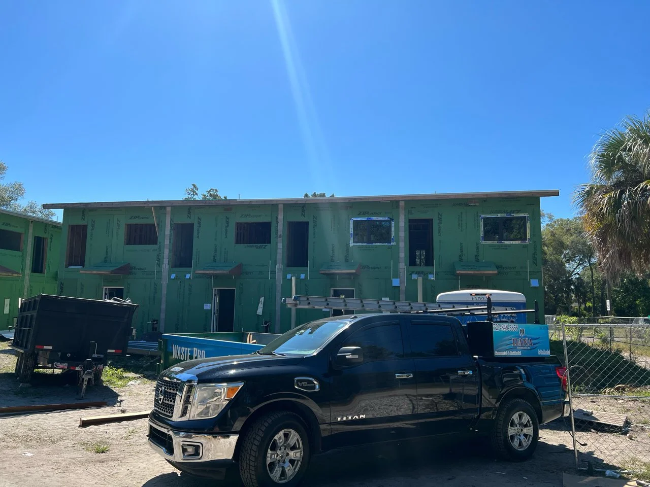 Construction site with a partially built green house, a black pickup truck parked in front, and construction equipment nearby under a clear blue sky.